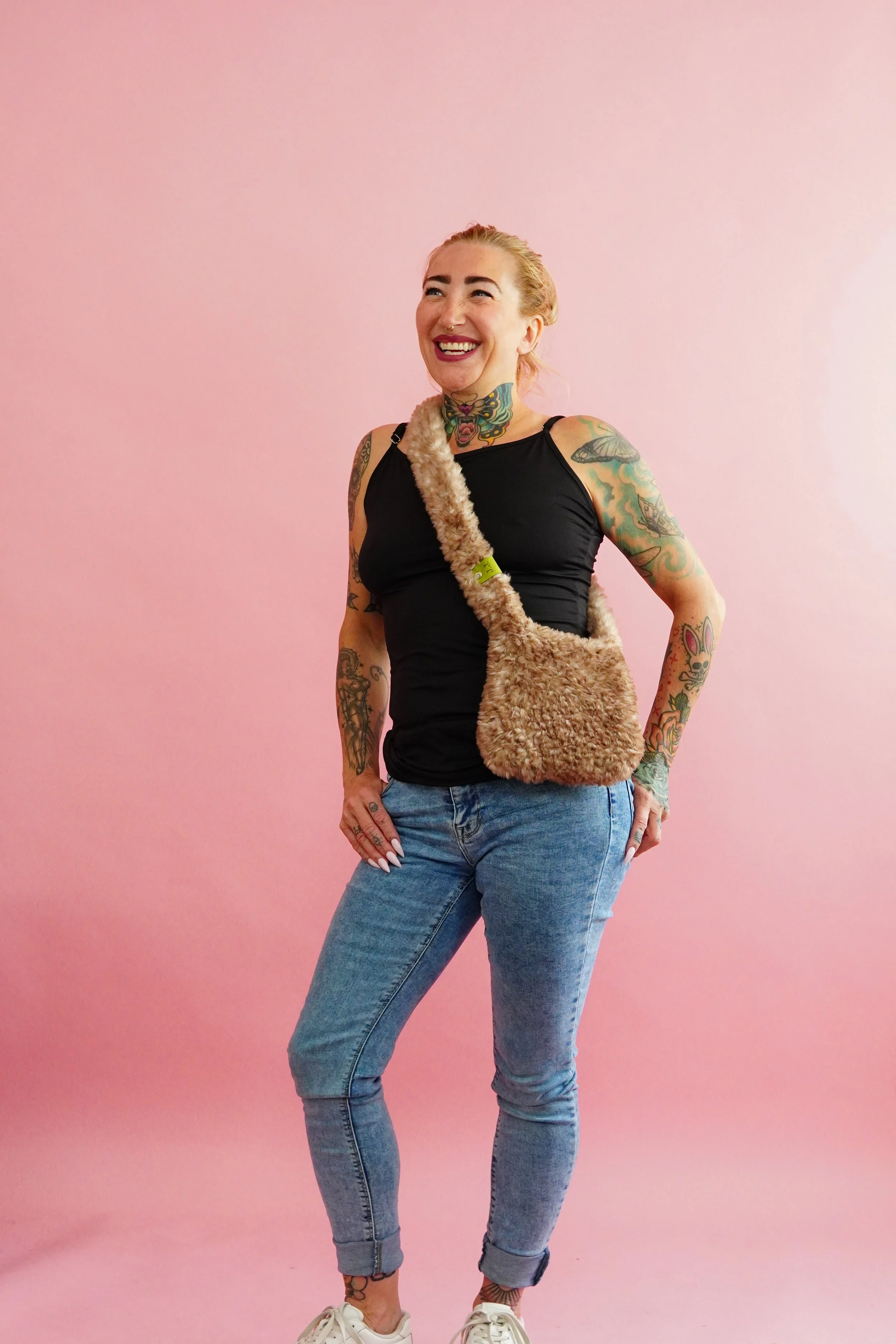 A woman with tattoos on her arms and neck, wearing a black tank top, light blue jeans, and white sneakers, stands against a pink background, smiling while carrying a fuzzy tan handmade crocheted shoulder bag.