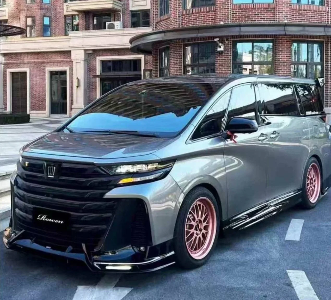 A silver minivan with custom pink wheels and black modifications parked on a city street in front of a brick building.