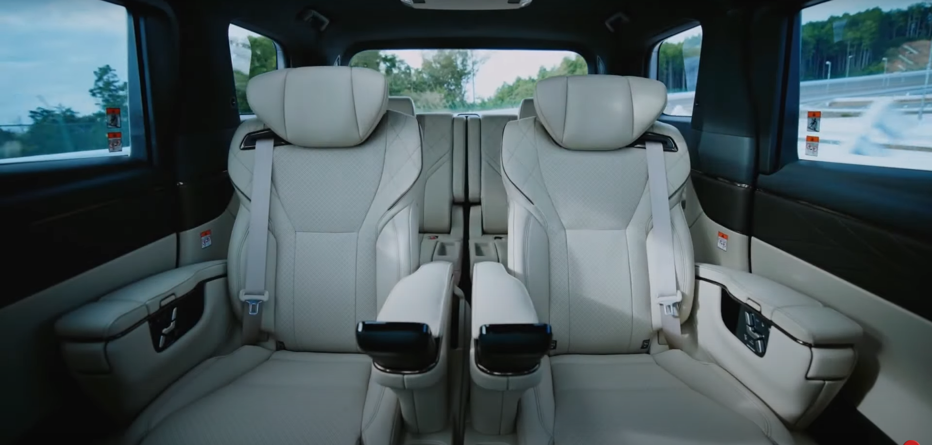 Interior of a luxury vehicle showing two beige leather captain's chairs with headrests, armrests, and seatbelts, with windows revealing a highway and greenery outside.