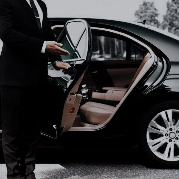Person in formal attire standing outside a black luxury car with an open front passenger door, holding a tablet.