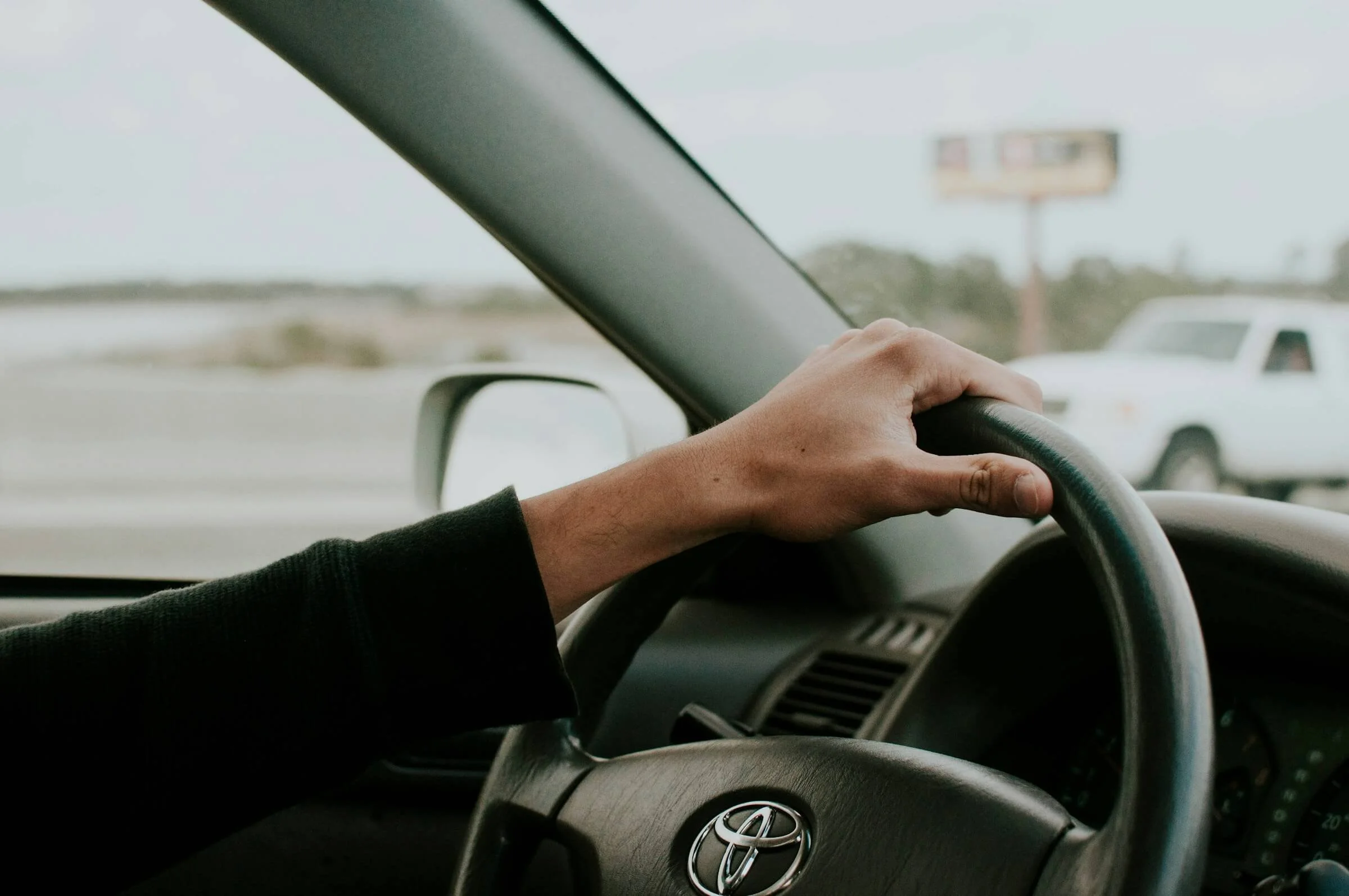 A person's hand on the steering wheel of a Toyota vehicle, driving on a highway with other cars visible outside.