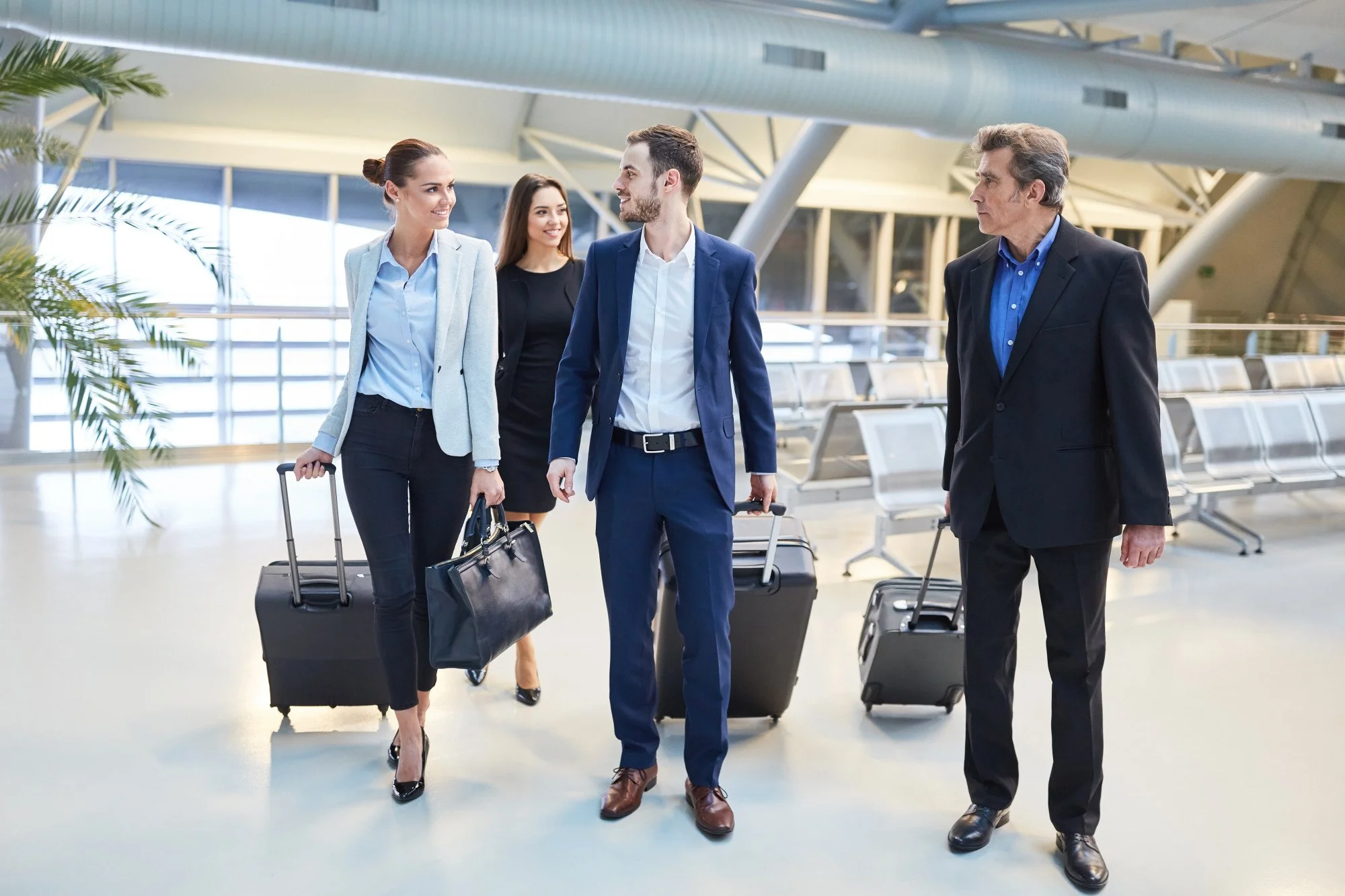 Four business professionals at an airport, walking with luggage, engaging in conversation.