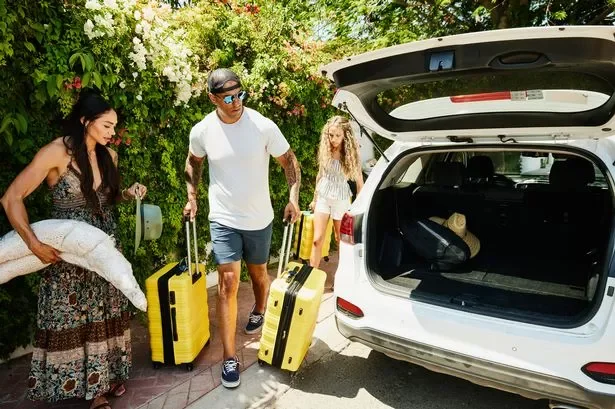 A family loading luggage into the trunk of a white SUV on a sunny day, surrounded by greenery.