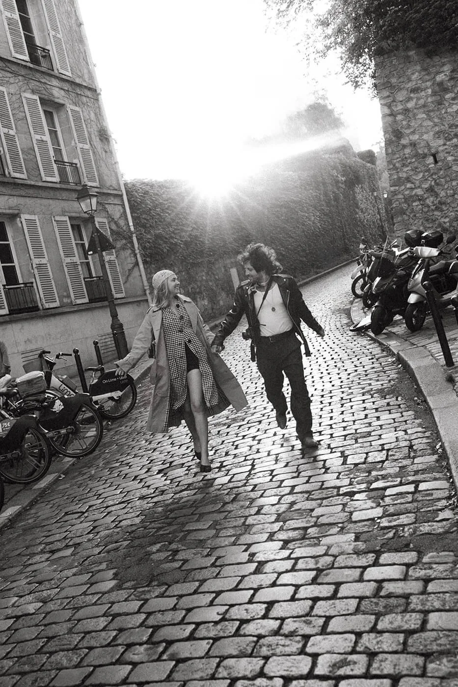 Black and white photo of a couple running hand in hand through a Paris street, cinematic engagement session in Montmartre.