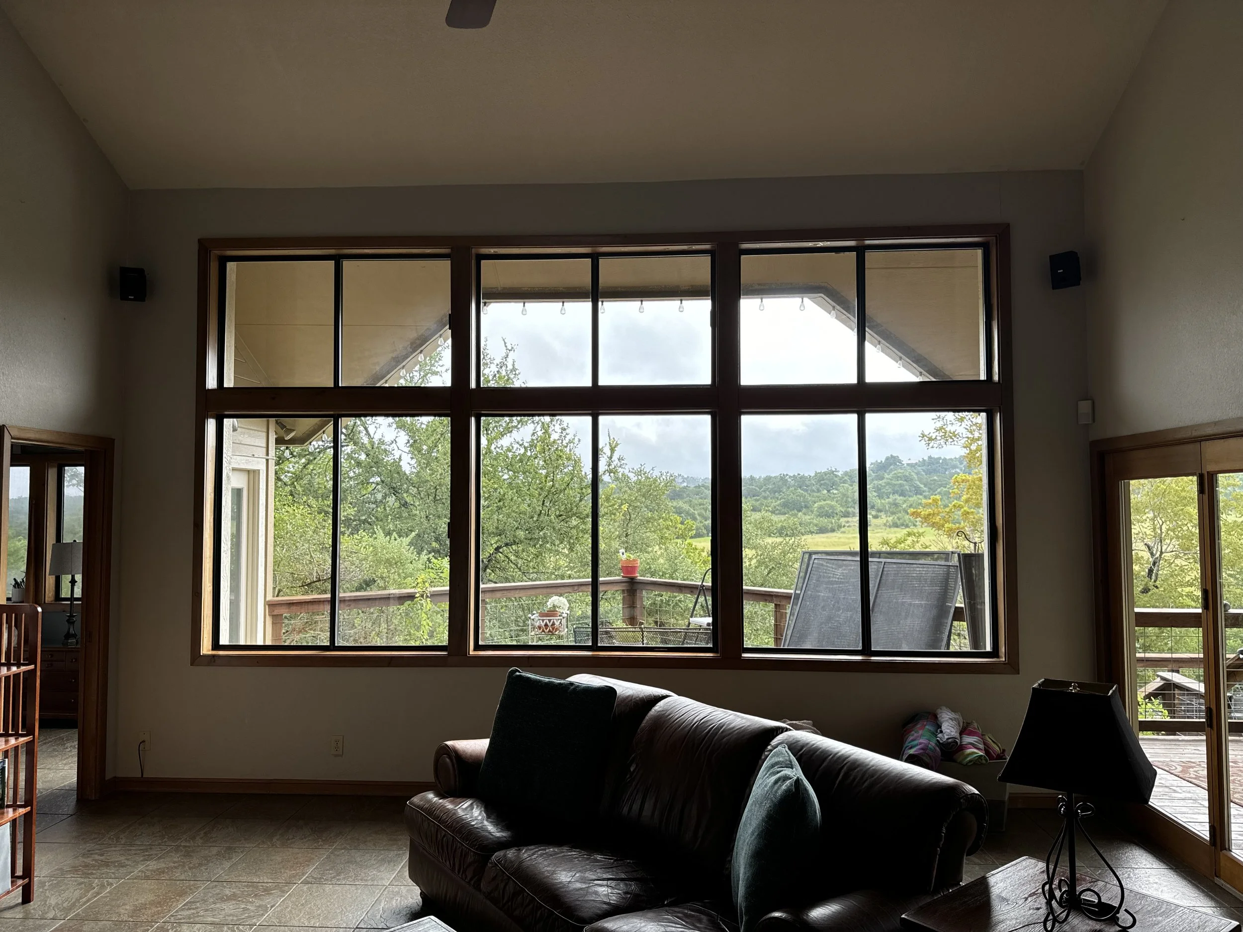 Living room with large front window showing a balcony, outdoor landscape, and cloudy sky, with a leather sofa, a side table with a lamp, and patio furniture outside.