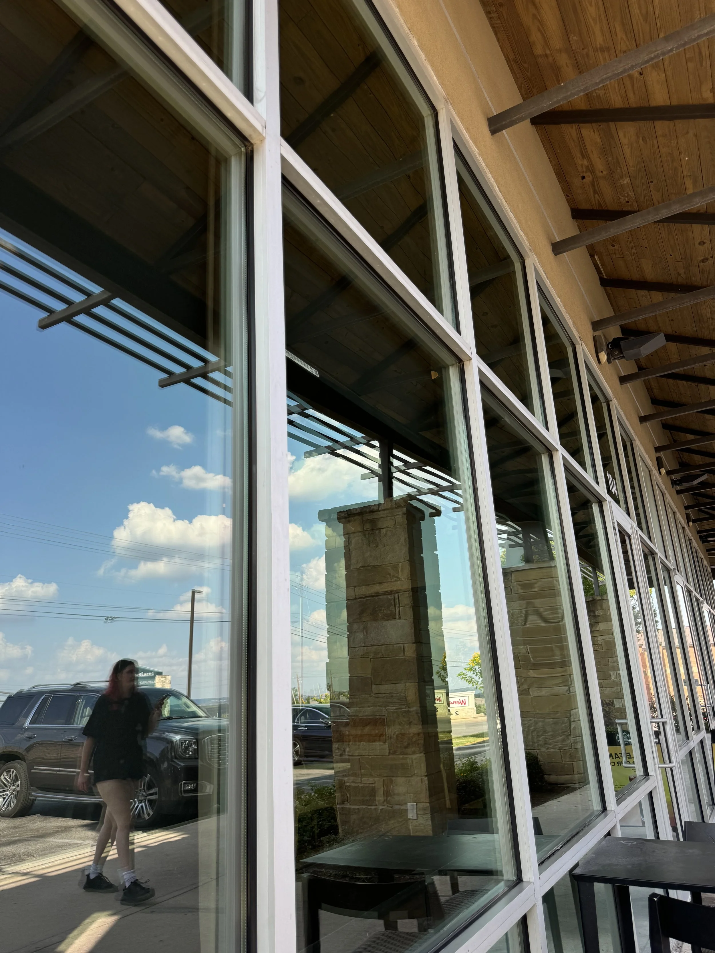 Reflection of a woman walking outside a building with large glass windows, parked cars, and cloudy sky visible in the windows.