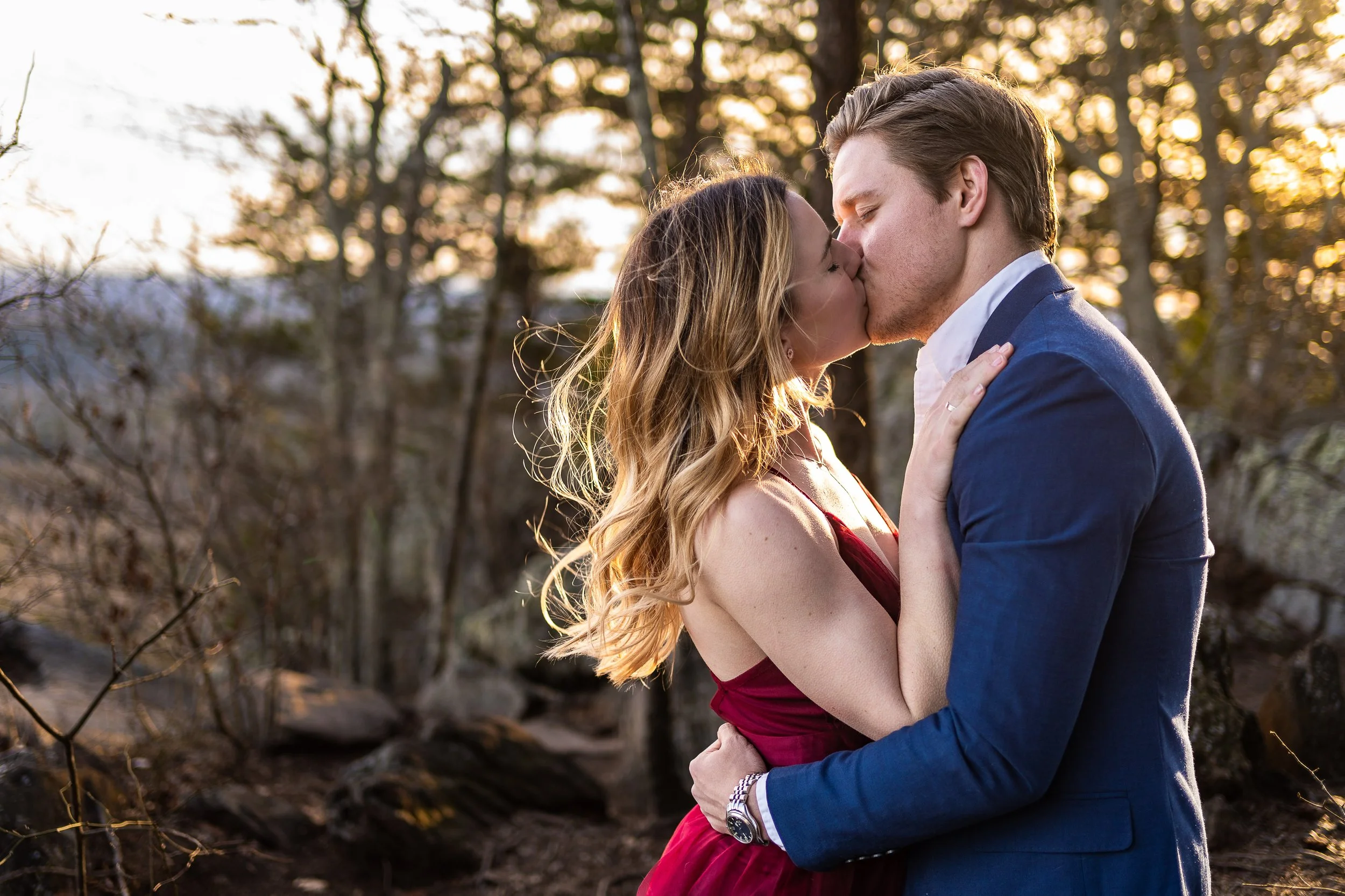 A couple kissing outdoors during sunset, with the woman wearing a red dress and the man in a blue suit.