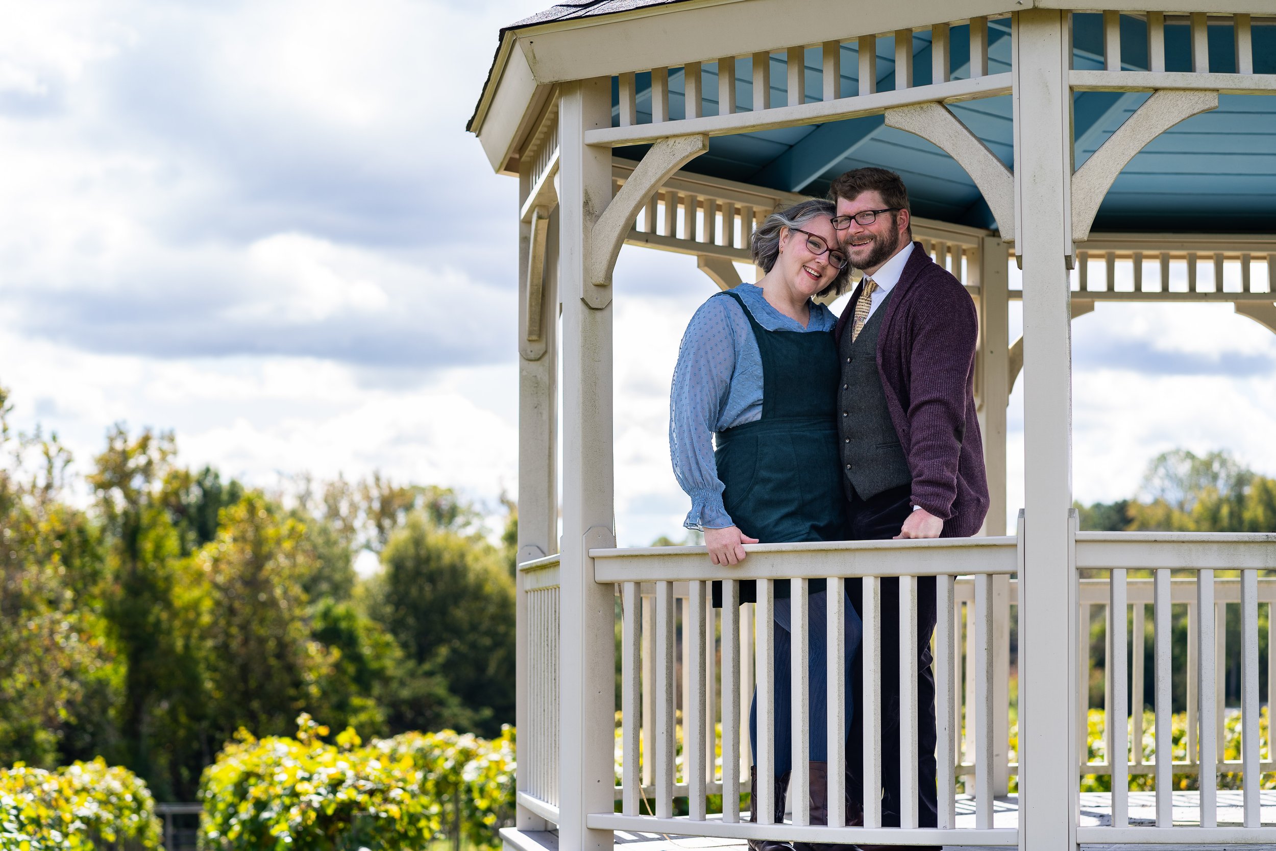 Two people, a woman with gray hair and glasses wearing a blue blouse and dark overalls, and a man with glasses and a beard wearing a white shirt, tie, gray vest, and maroon cardigan, standing close together on a gazebo balcony outdoors, smiling.