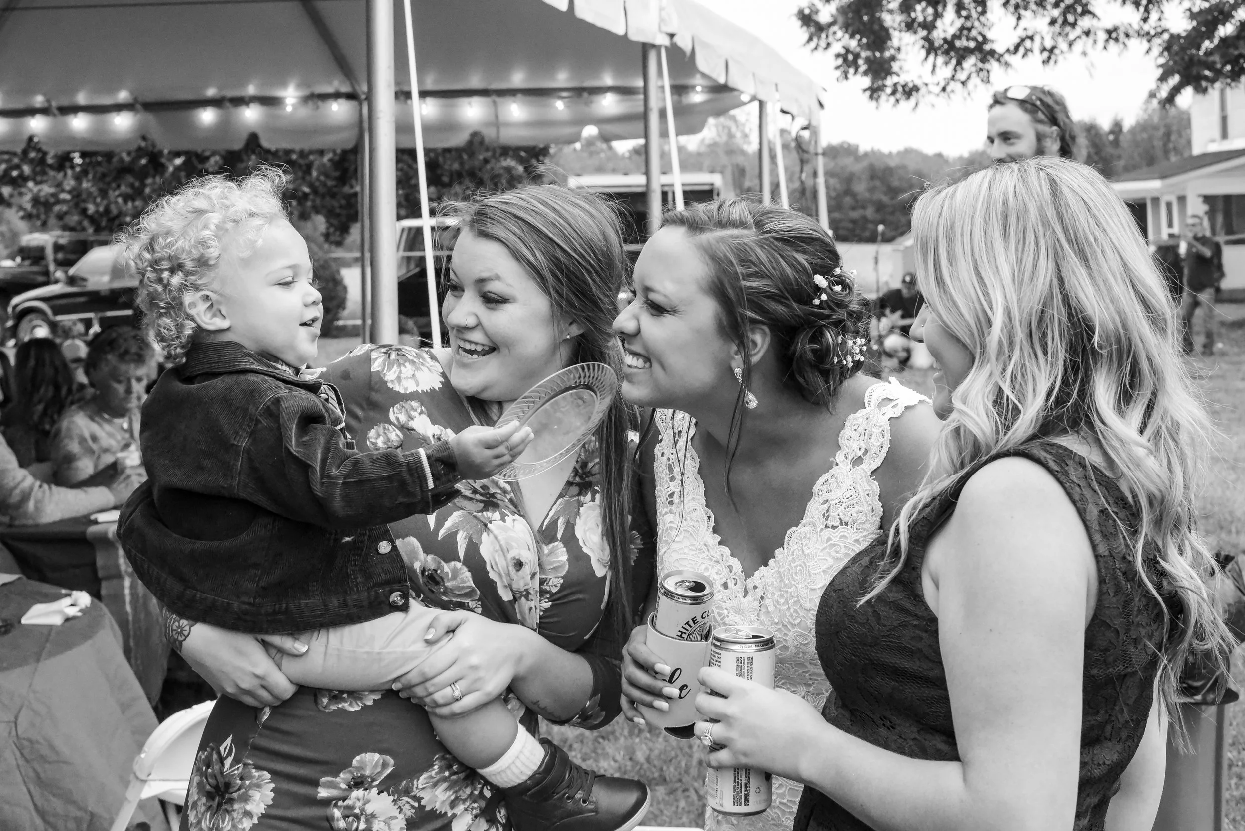 Three women and a young child are smiling and interacting at an outdoor gathering under a canopy tent, holding drinks and a plate of food.