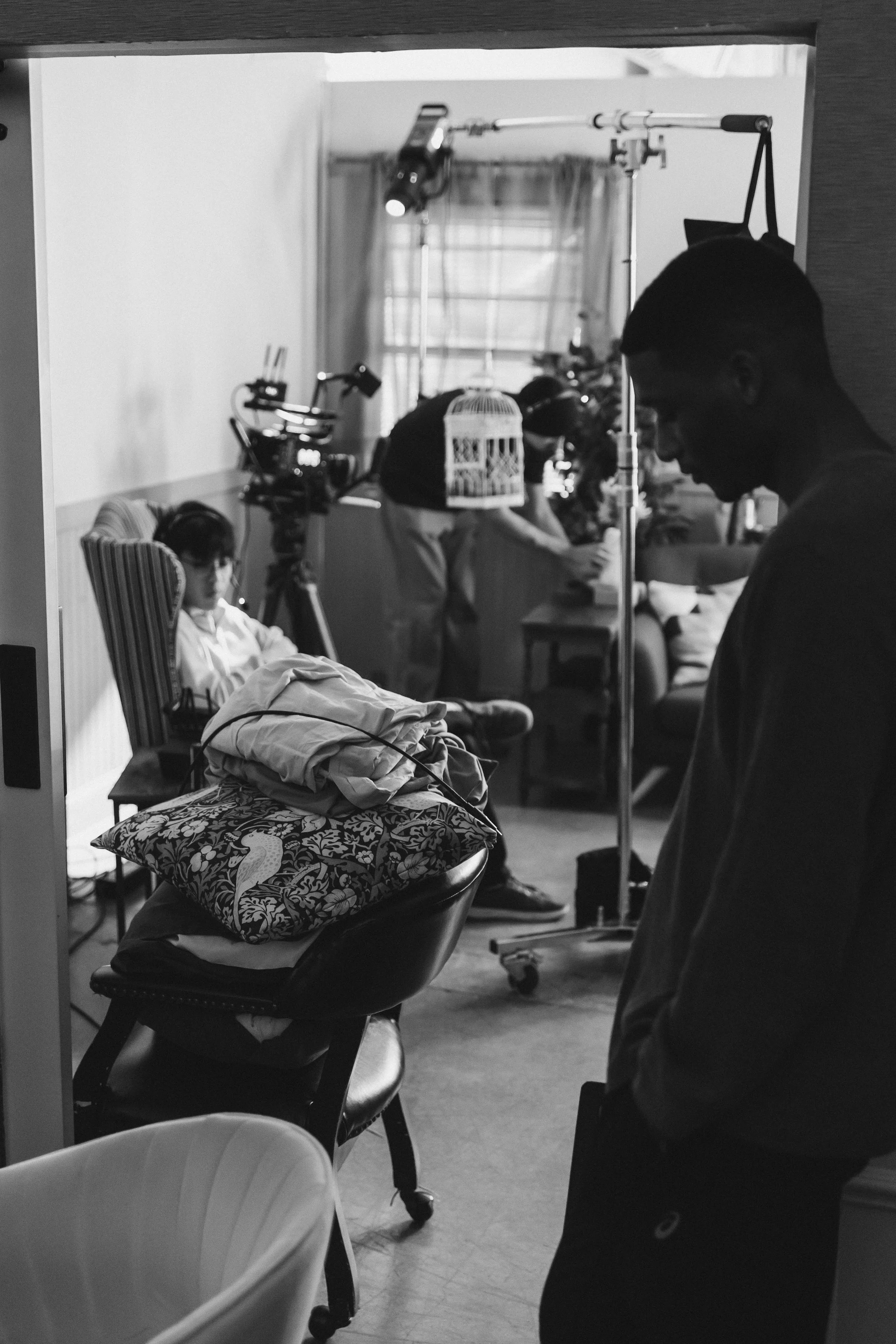 Black and white photo of a film set with a young girl sitting in a striped armchair, a camera on a tripod, and crew members preparing equipment, viewed through a window.