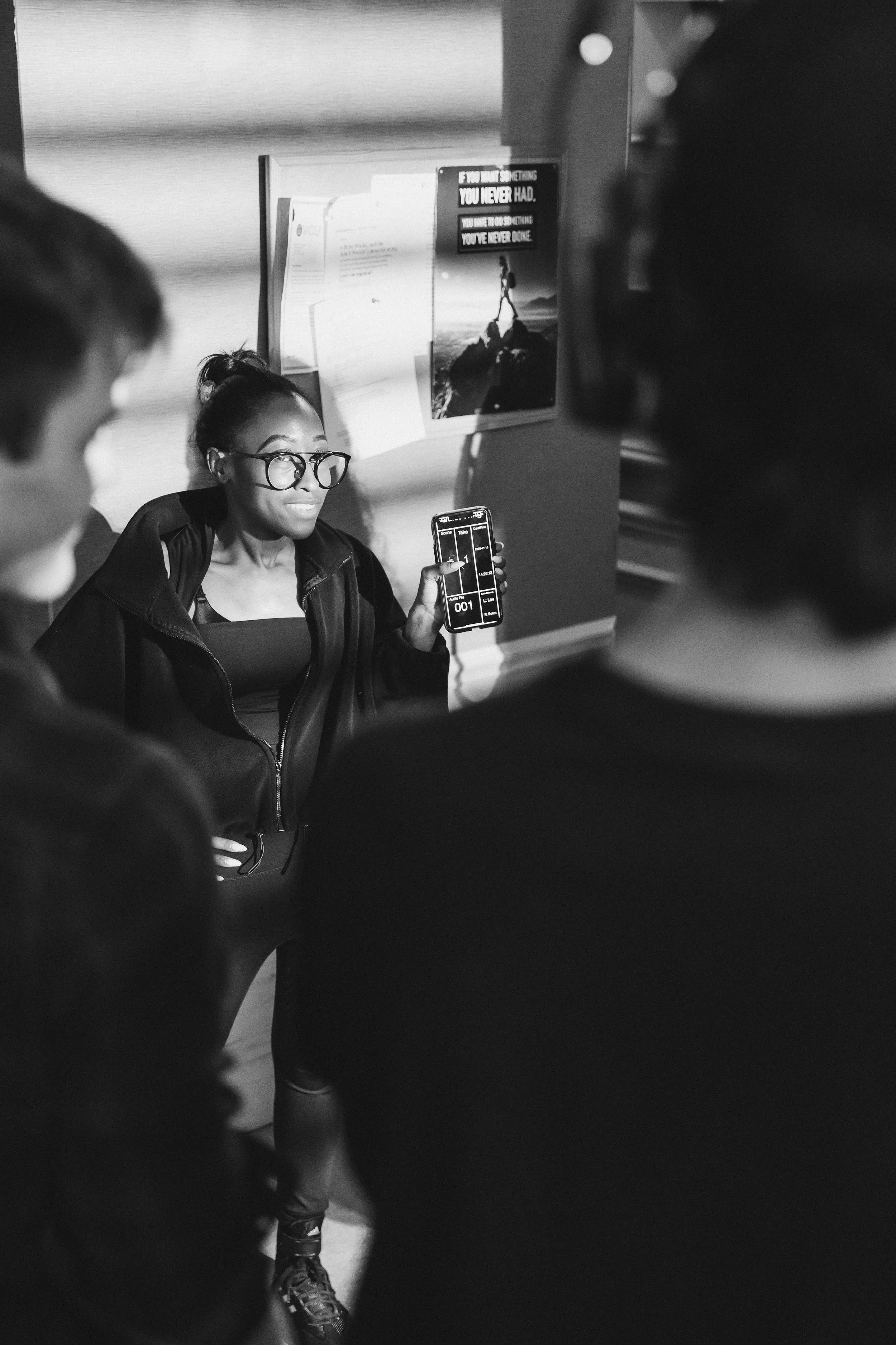 A woman sitting on the floor holding a smartphone with a timer app, surrounded by a group of people in a public indoor space, with posters on the wall in the background.