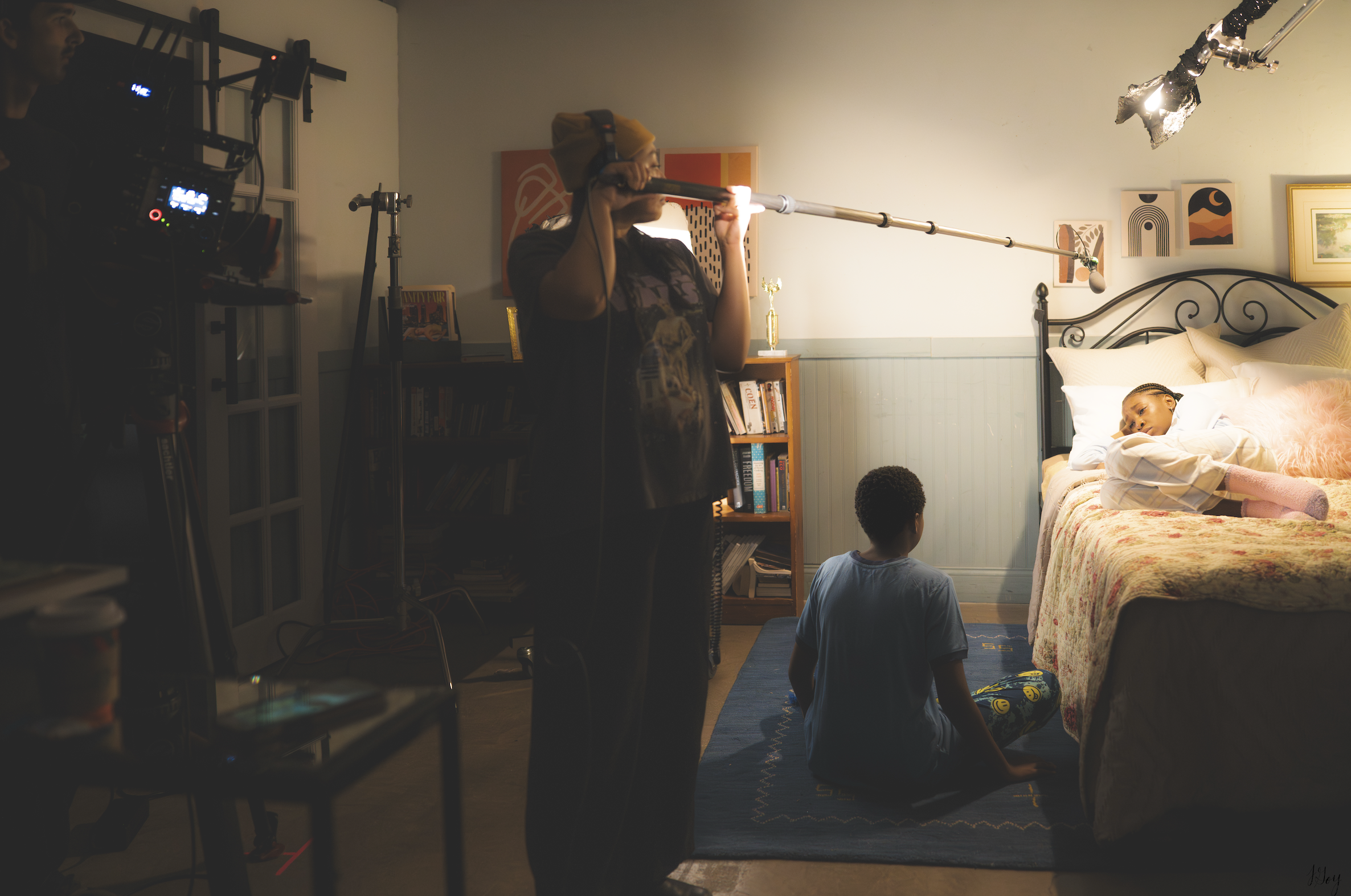 A person holding a boom microphone operates it above a girl lying on a bed, with a boy sitting on the floor facing the girl in a dimly lit room with artwork and books.