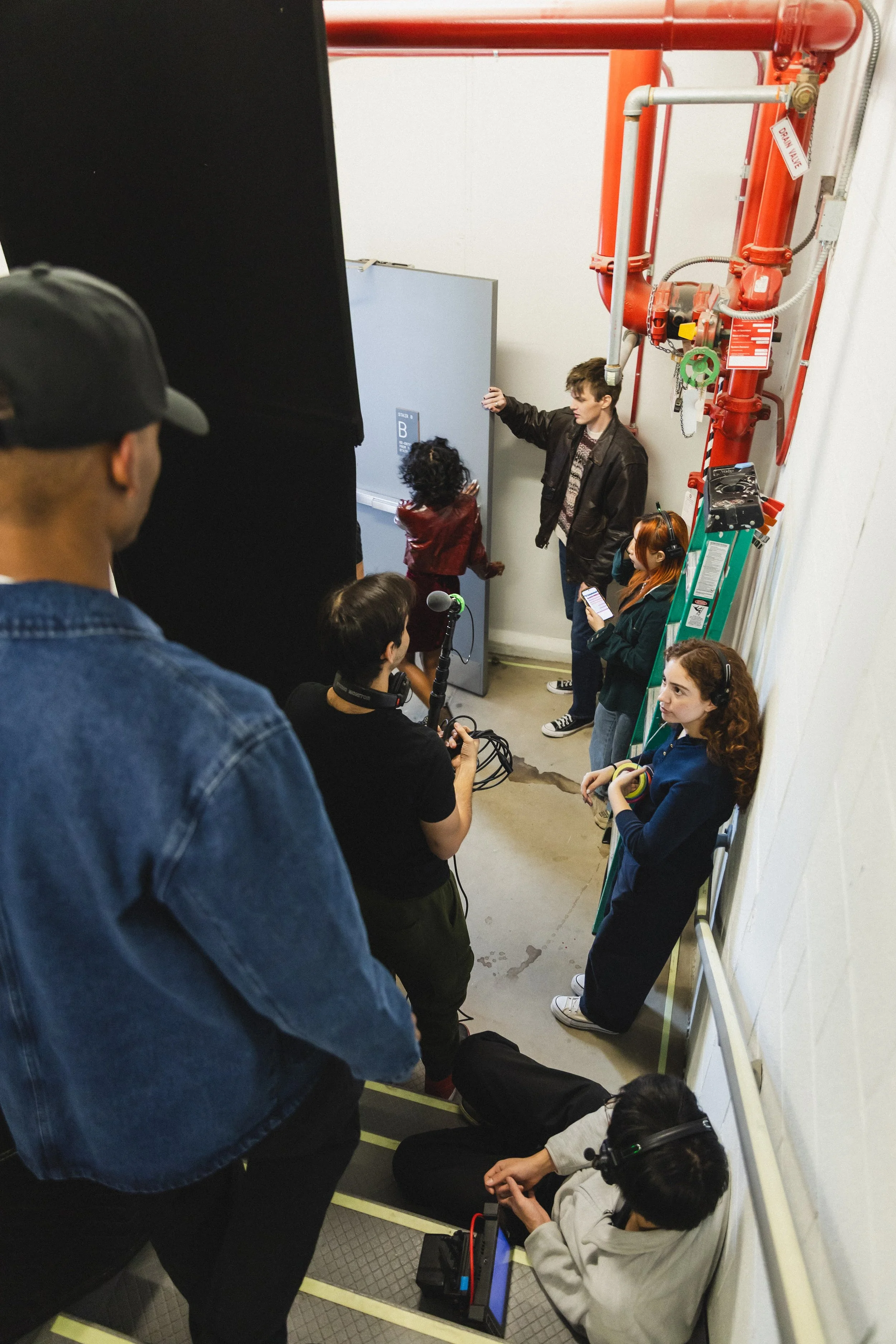 Group of people, some wearing headphones and holding equipment, in a narrow space with a ladder, pipes, and industrial equipment.