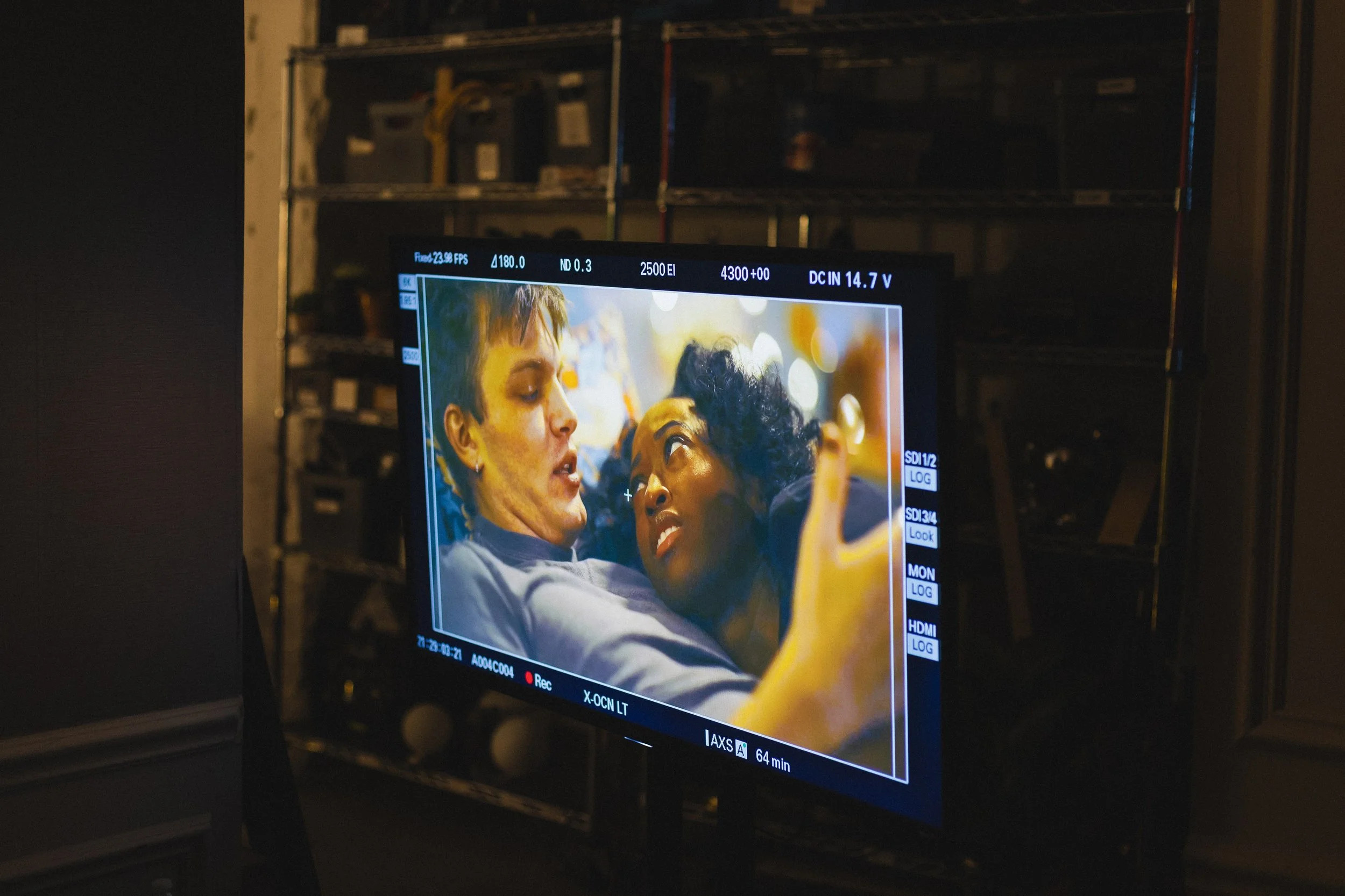 A scene from a film shot on a camera monitor shows two women lying close together, looking into each other's eyes, with one woman holding her hand near her face. The background is blurred, with warm lighting.