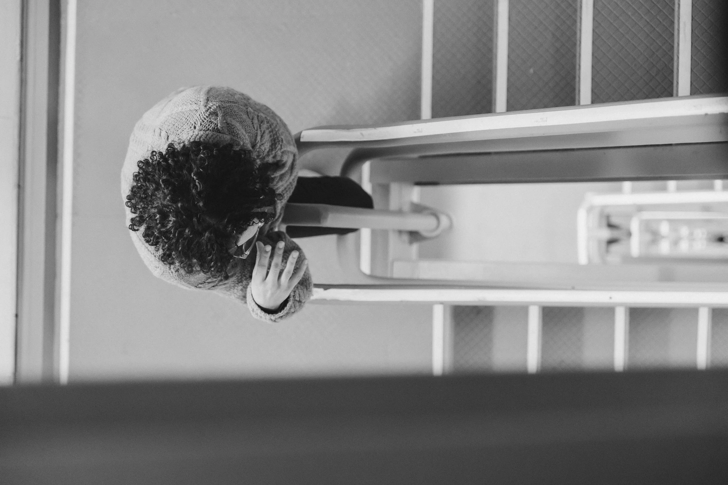 A child with curly hair wearing glasses and a knitted hat, resting their chin on their hand while sitting at a desk or table, looking downward.