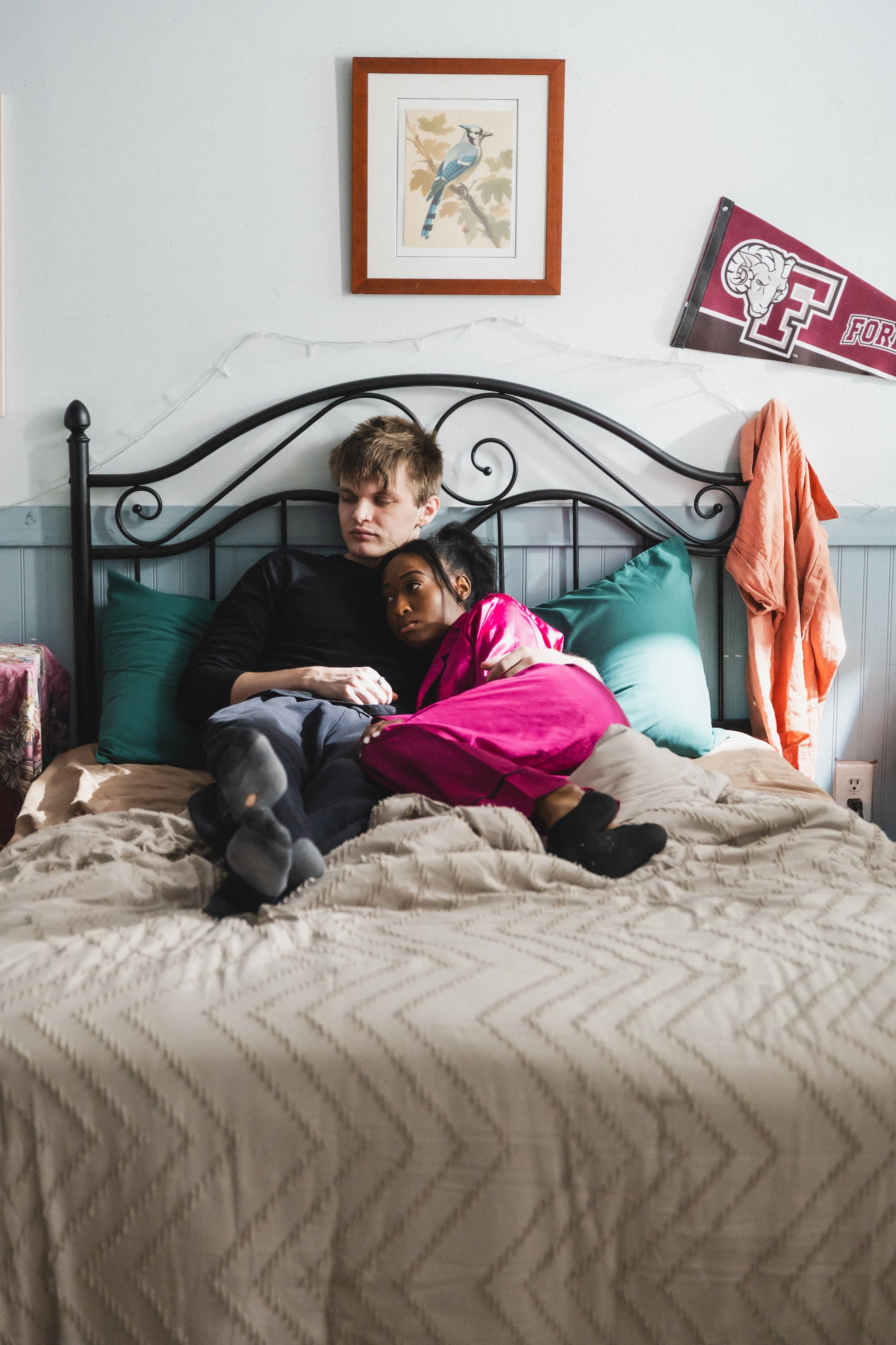A young man and woman lie on a bed, with the woman resting her head on the man's chest, in a room with artwork and college banners on the wall.