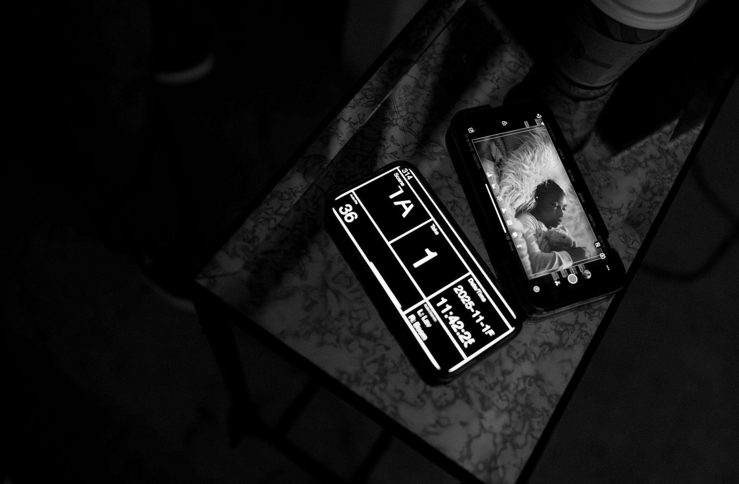 A smartphone displaying a black and white photo of a woman with long hair lying on a pillow, holding a pillow close, with an ice cream cone on the table next to it. The phone is on a marble tabletop with a ticket and a coffee cup nearby.