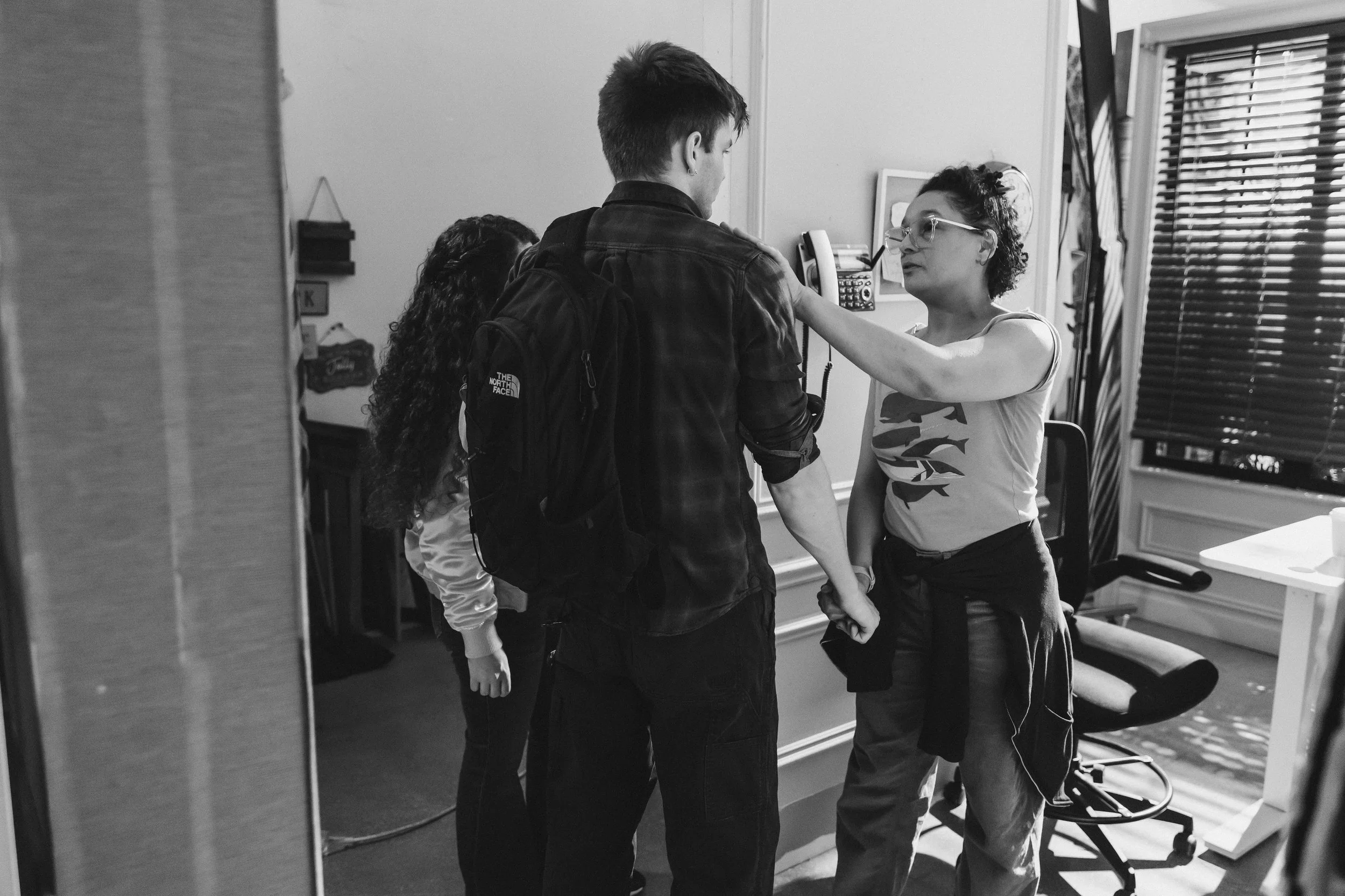 A woman with glasses and a tattoo on her arm is speaking to a young man and two women in a room with office furniture, while touching the man's shoulder.
