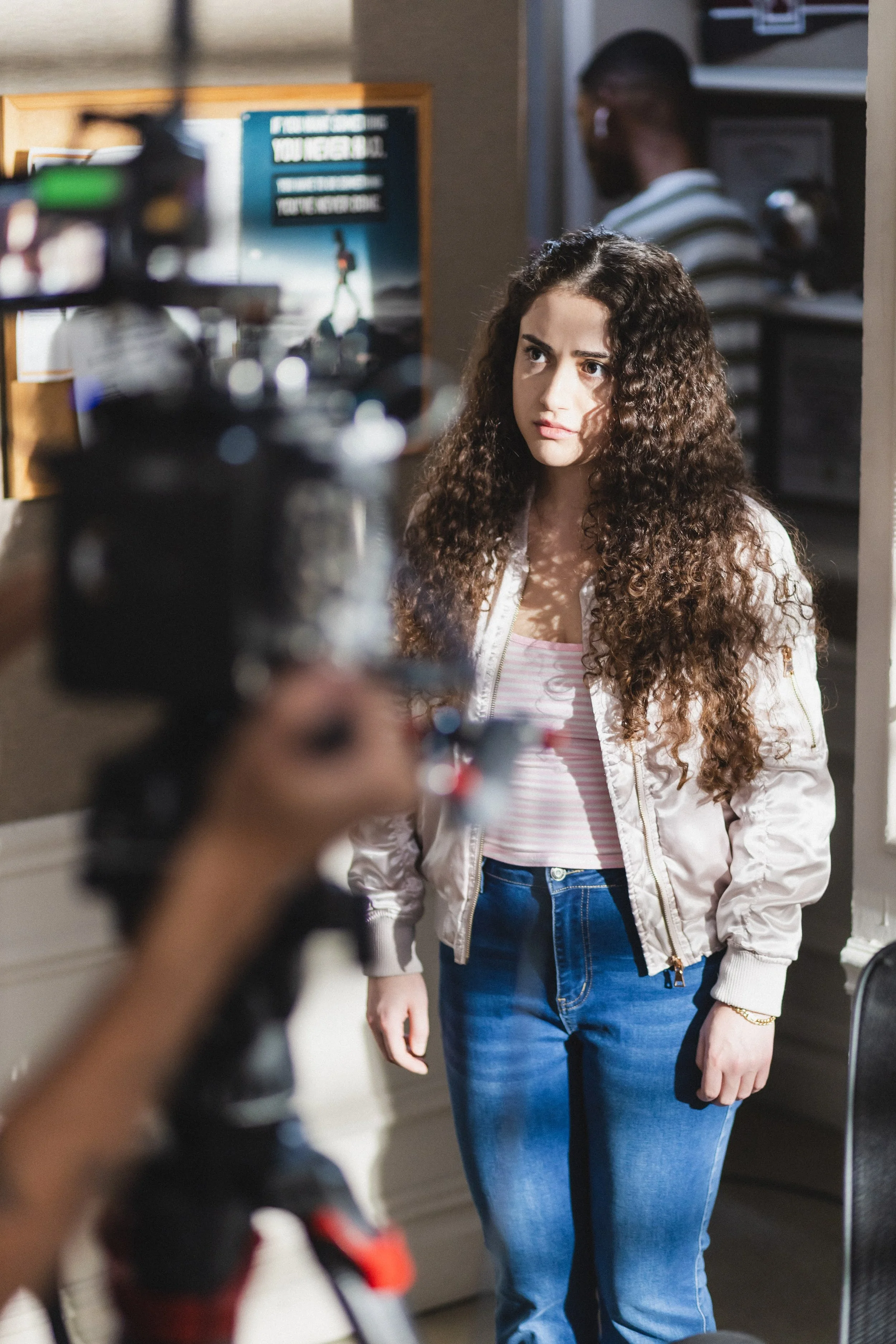 A young woman with curly hair looks attentively at the camera during a video recording, standing in a room with a bulletin board and another person in the background.