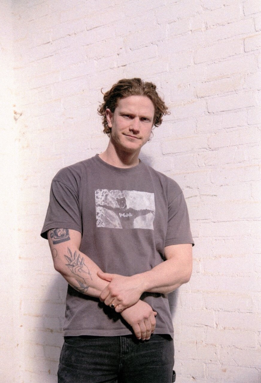 A young man with curly hair and tattoos on his arm, standing against a white brick wall, wearing a gray t-shirt with a graphic print and black jeans.