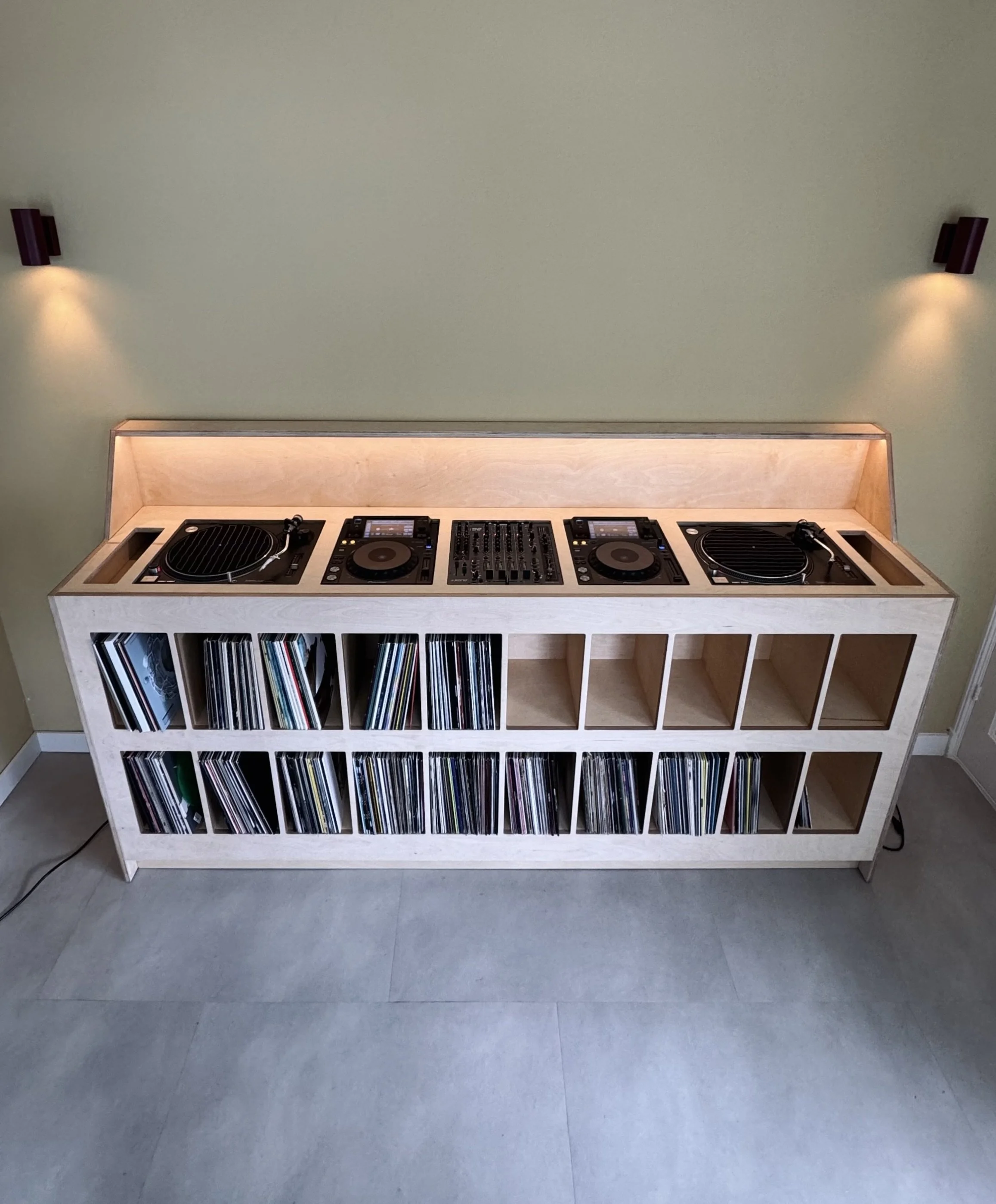 Wooden DJ station with turntables, mixer, and vinyl records on a shelving unit against a green wall with wall sconces.