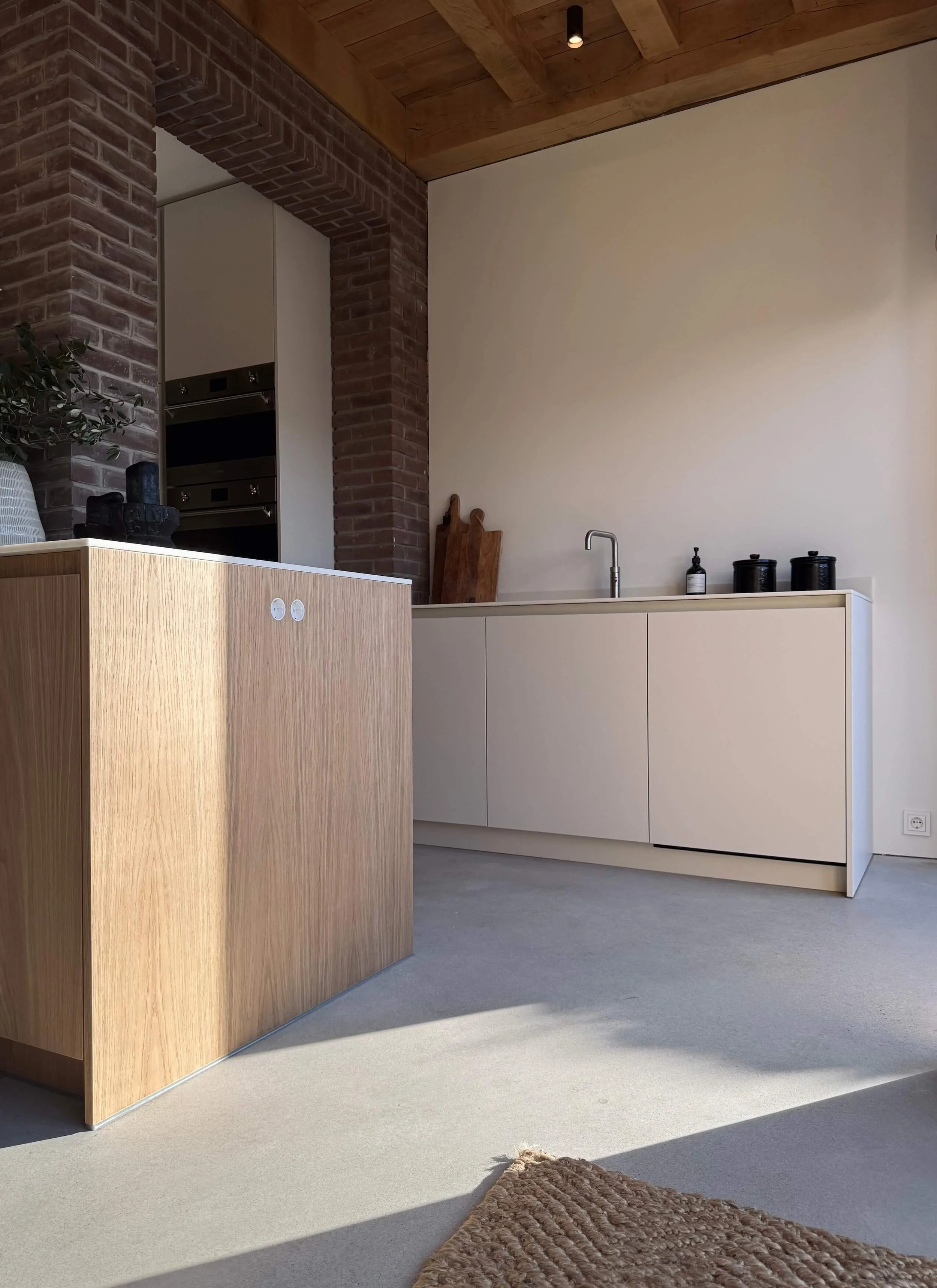 Modern kitchen with white cabinetry, a white countertop, and a stainless steel faucet. A wooden island with a light finish is in the foreground, and a bare wall with a wooden ceiling and exposed brick arch is visible in the background. Several black containers and wooden cutting boards are on the countertop.
