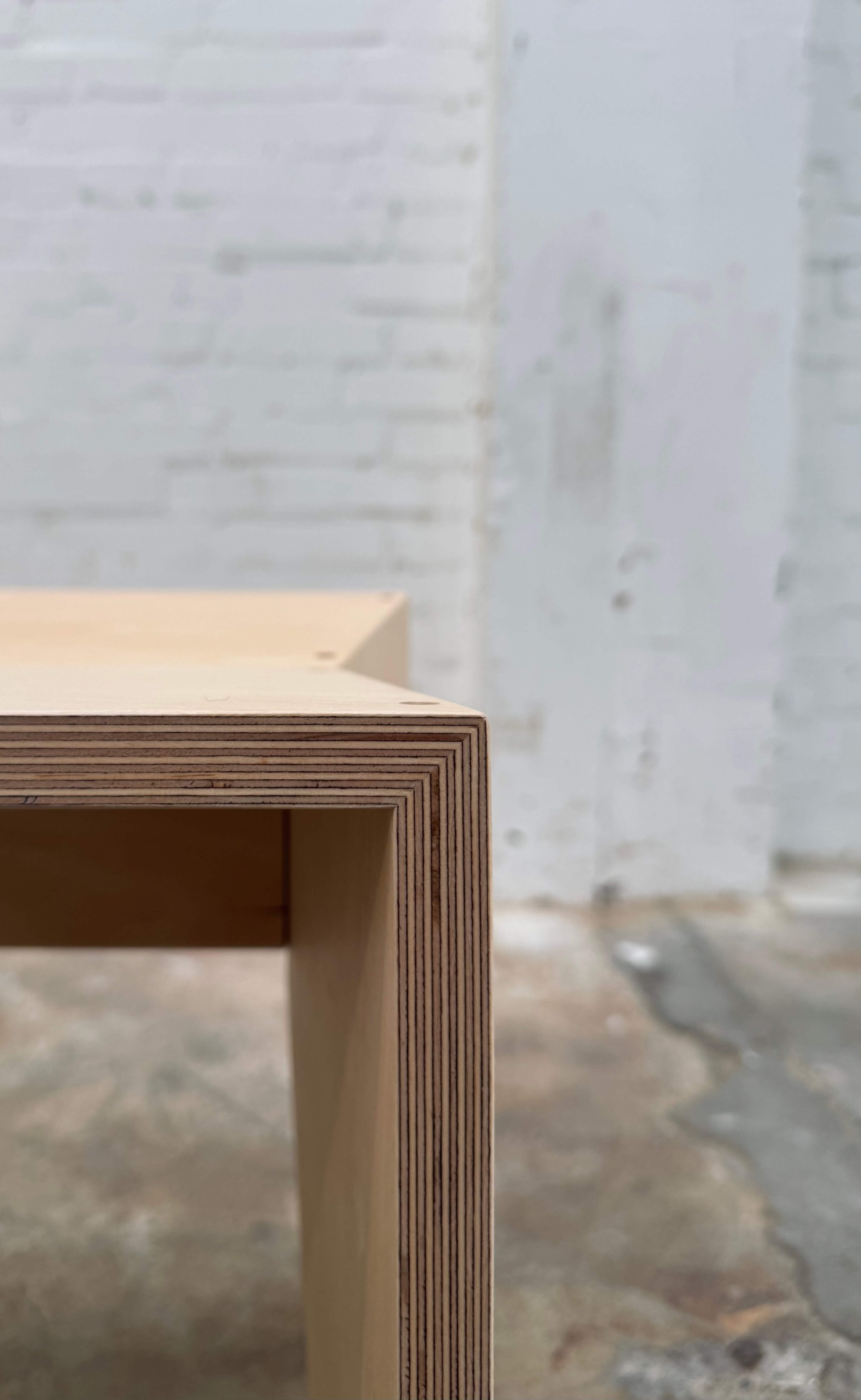 Close-up of a wooden piece of furniture with a layered edge, set against a blurred background of white brick wall and concrete floor.