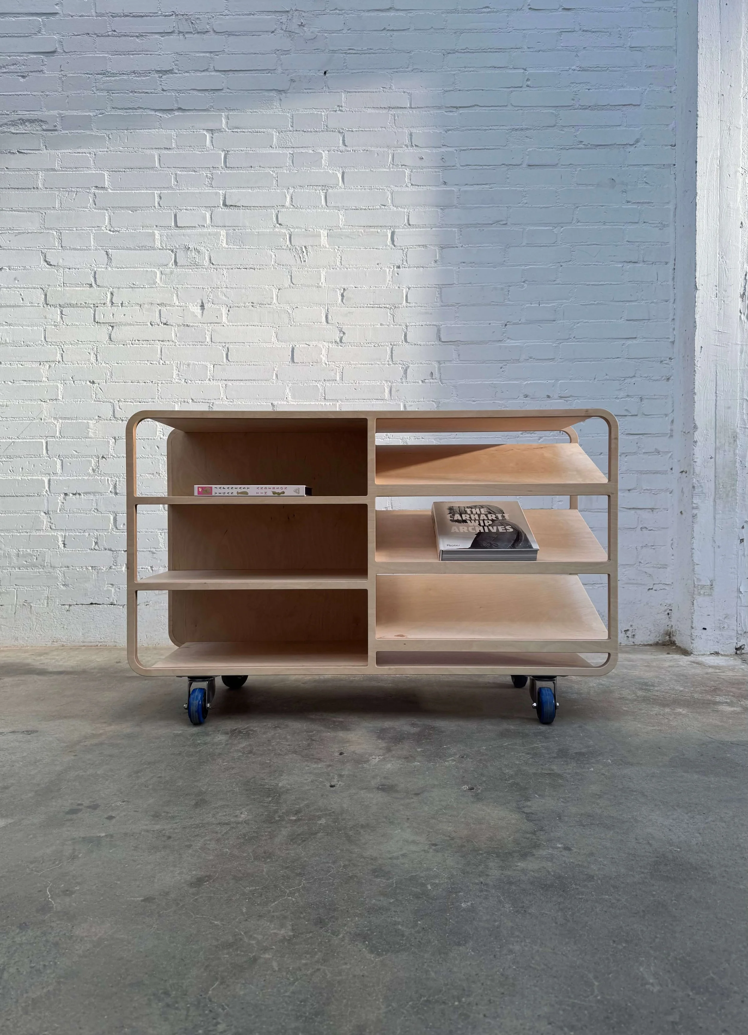 A wooden rolling cart with open shelves, placed on a concrete floor against a white brick wall. The cart has books on it, including one titled 'The Earth Archives'.