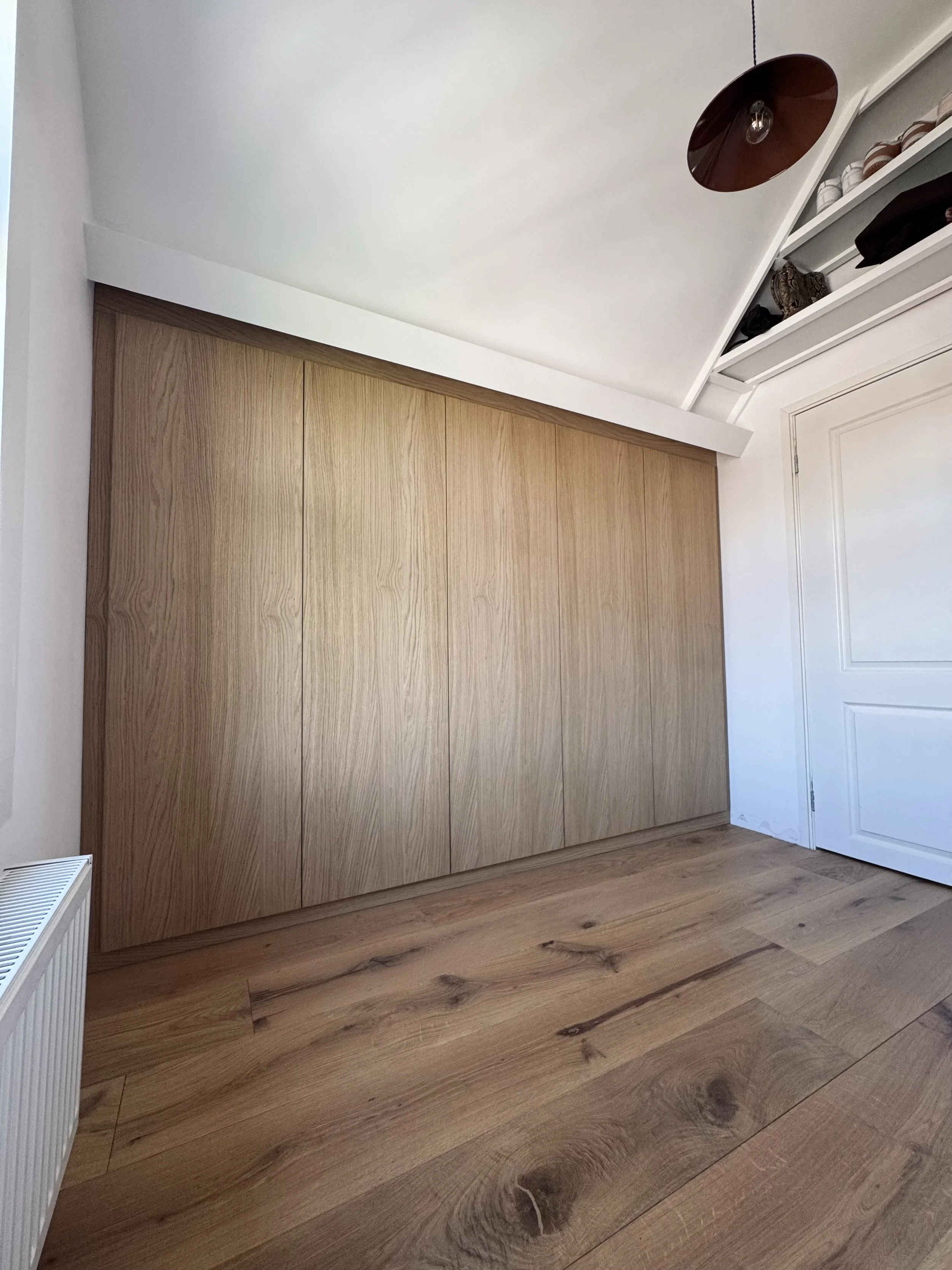 Room with wooden floor, large wooden closet, white walls, white door, and ceiling with a copper-colored hanging light