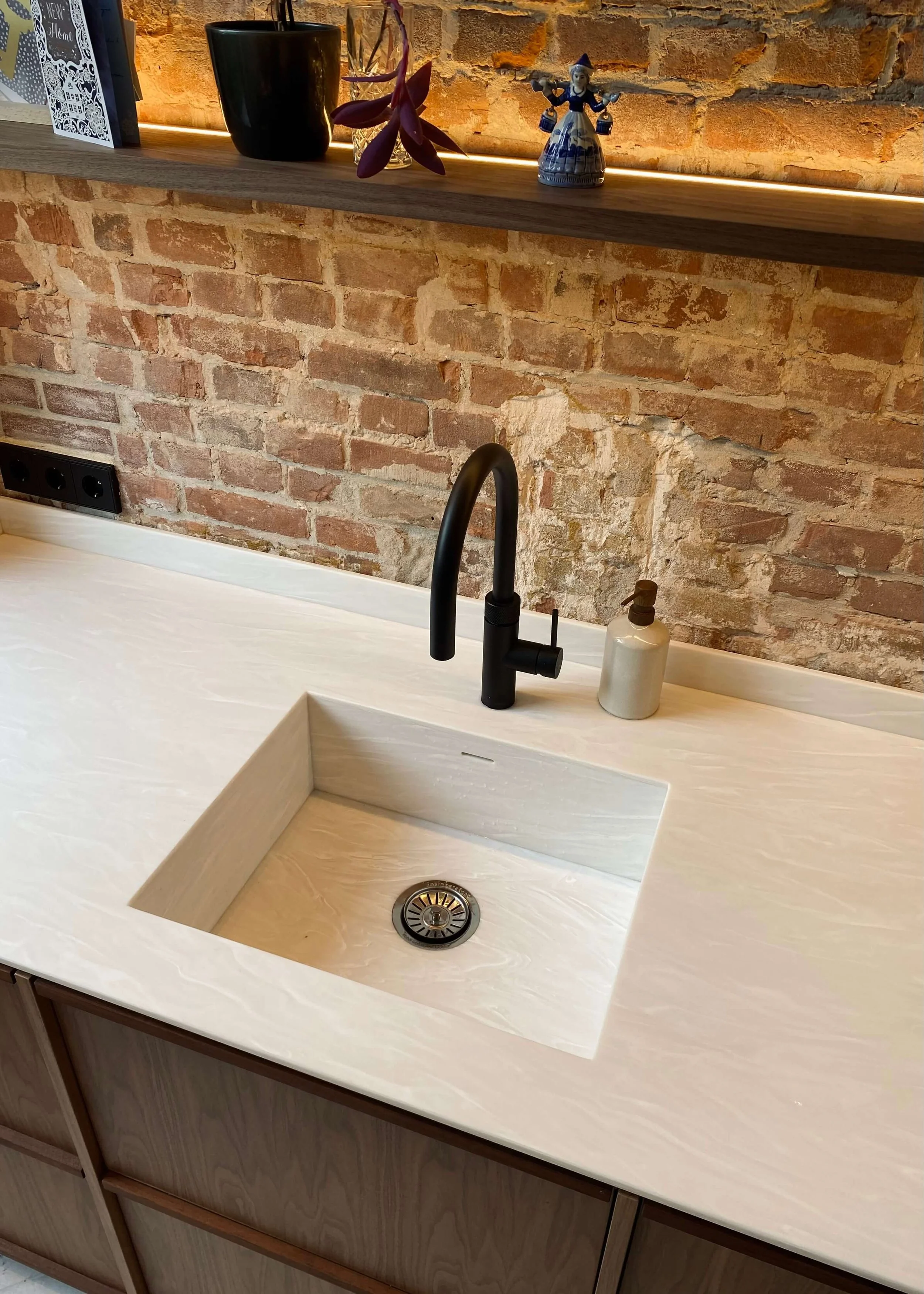 A modern kitchen sink with a black faucet and a white soap dispenser mounted on a white marble countertop, with an exposed brick wall and a wooden shelf with decorative items above.