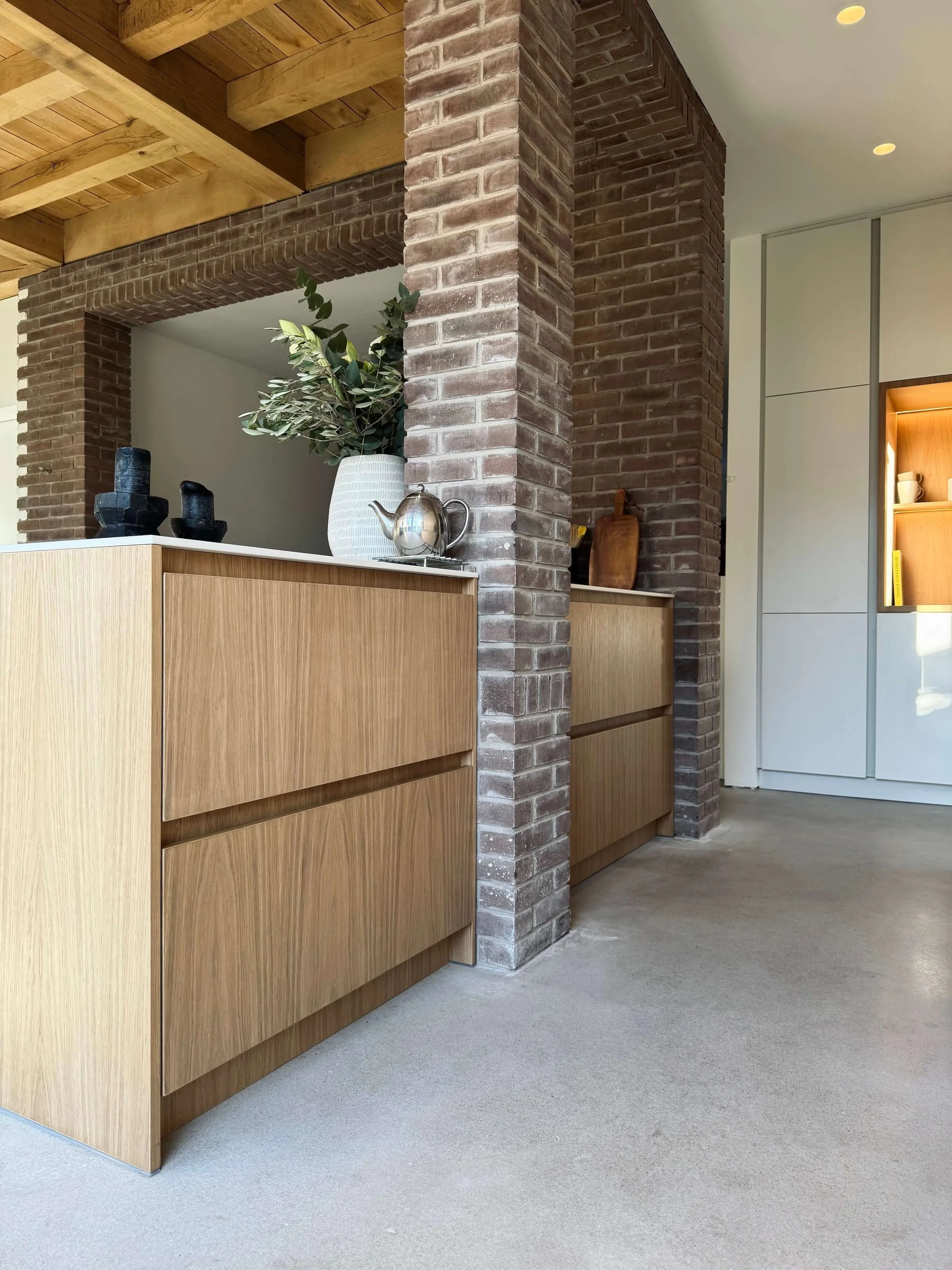 Modern interior with wooden cabinets, brick columns, a white vase with greenery, a teapot, and decorative objects, featuring a wooden ceiling and white walls.