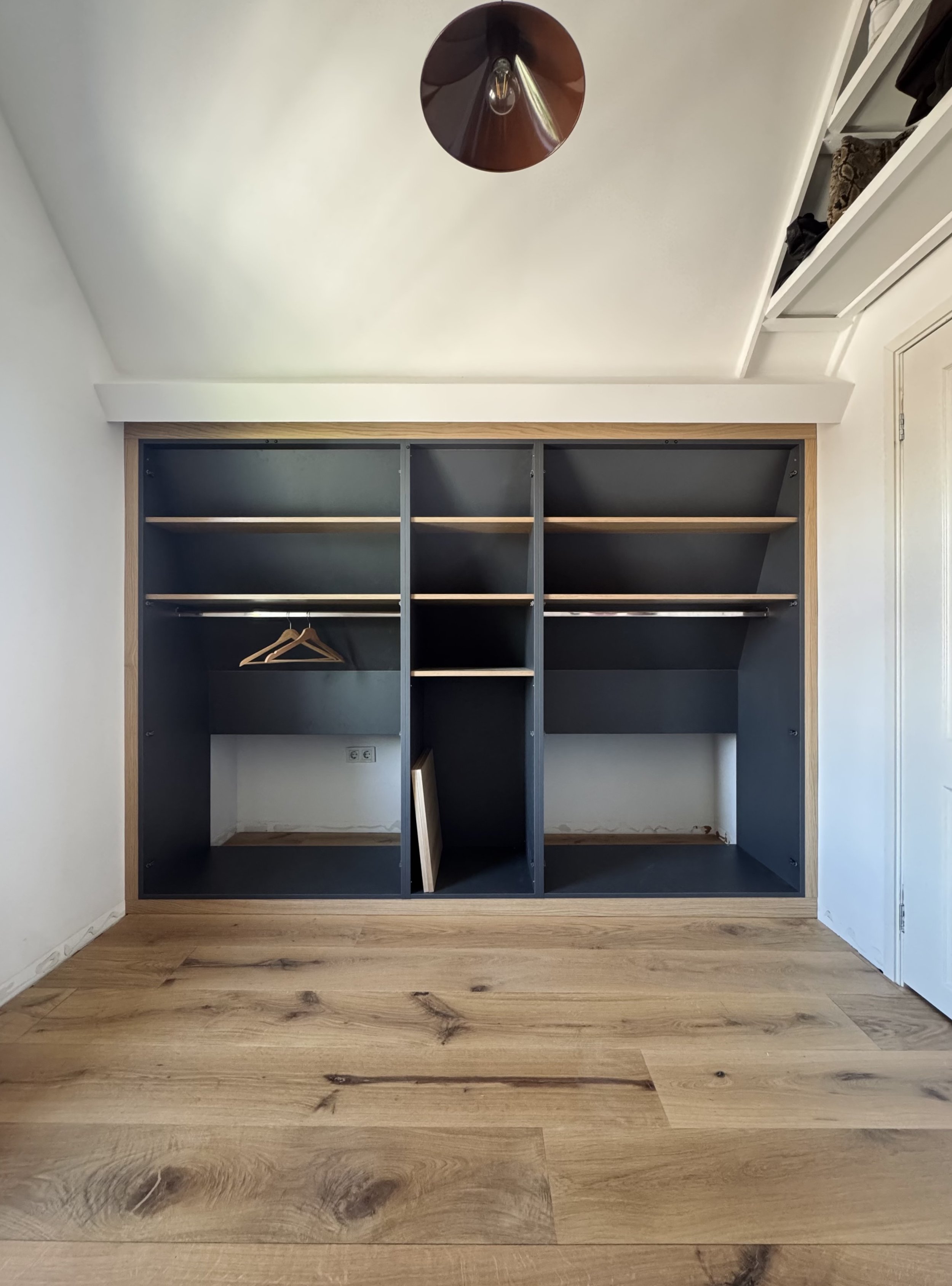 Empty closet with dark interior, wooden shelves, and a few hangers, set against a white wall with wood flooring.