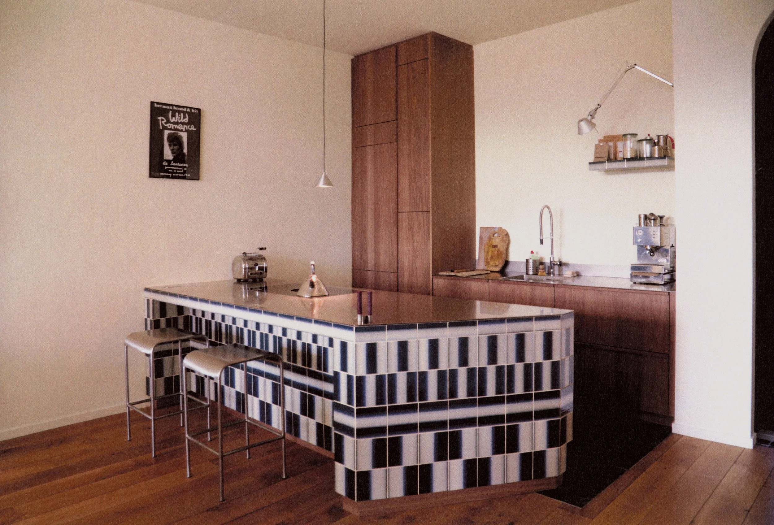 Modern kitchen with a tiled island, wooden cabinets, a sink, and a countertop with small appliances, decorated with a small shelf and framed artwork.
