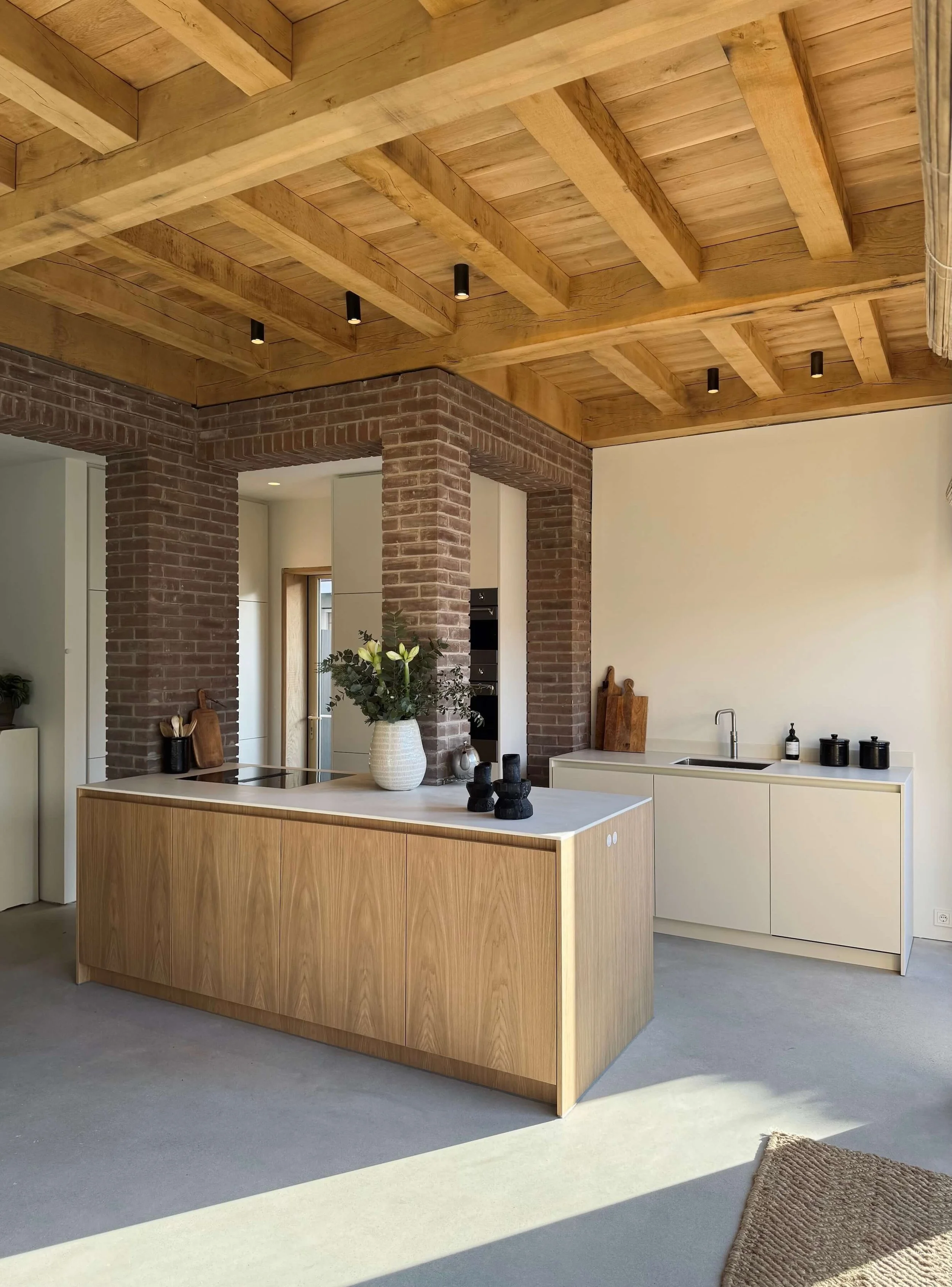 Modern kitchen with wooden ceiling, brick pillars, white cabinets, and a kitchen island with a vase of flowers.
