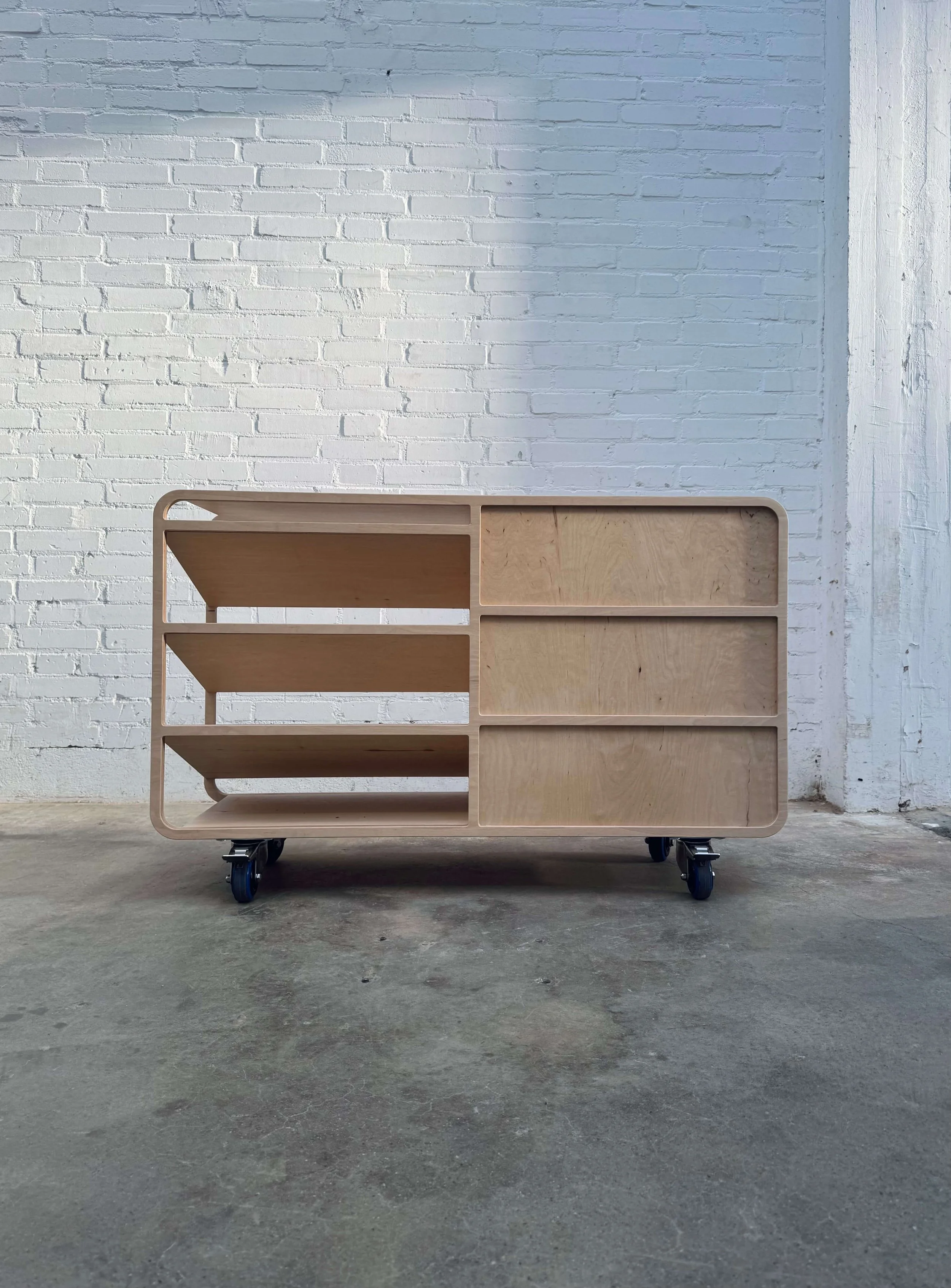A wooden mobile shelving unit with wheels, placed against a white brick wall on a concrete floor.