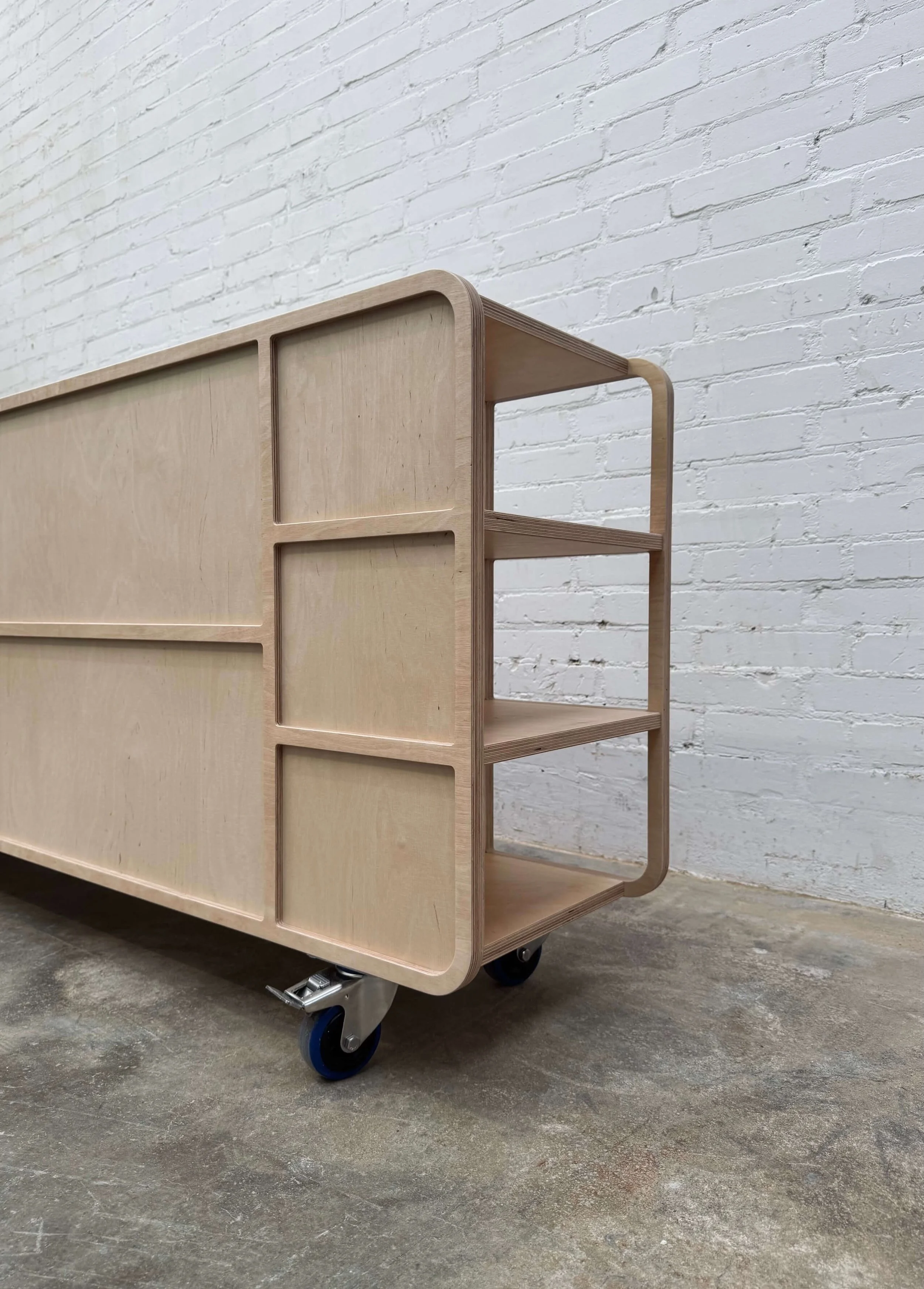Light-colored wooden storage cart with open shelving on four casters, positioned against a white brick wall.