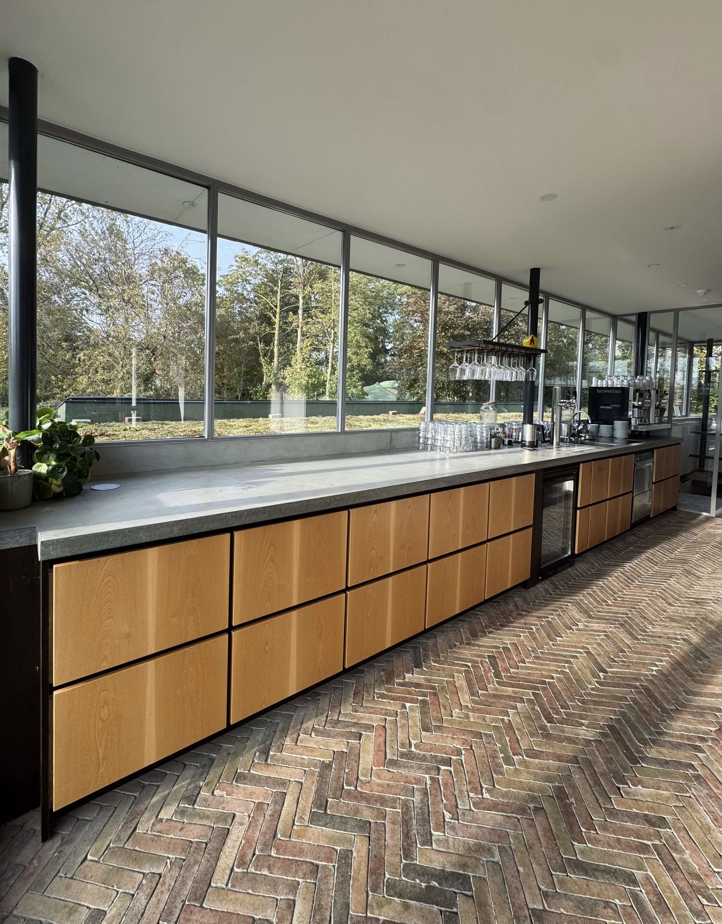 A modern coffee bar with a gray countertop, wooden cabinets, and a large window with a view of trees outside, featuring a coffee machine, glasses, and plants.