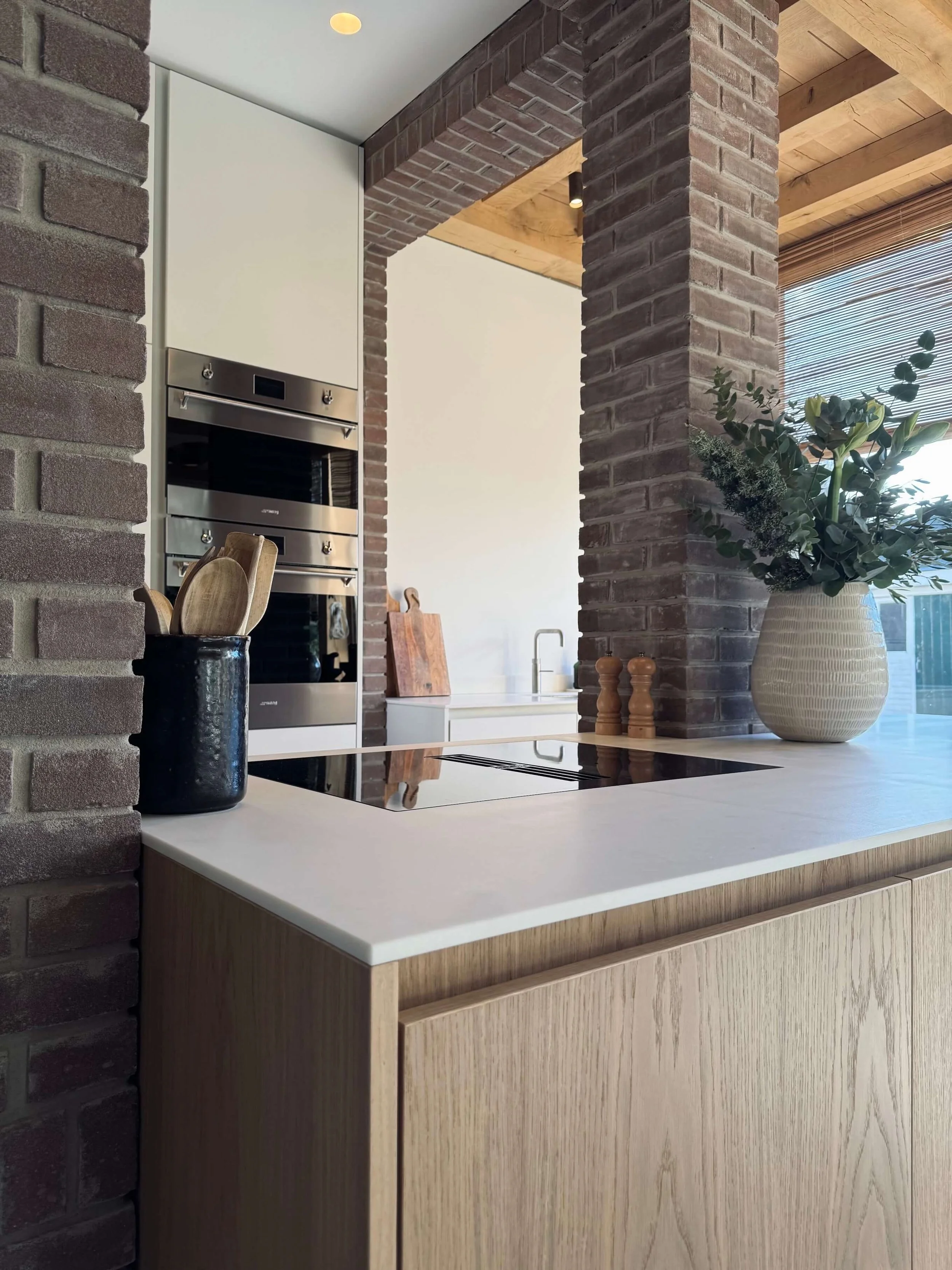 Modern kitchen with brick pillars, white cabinets, a built-in oven, a wooden countertop, and a large vase with green foliage.