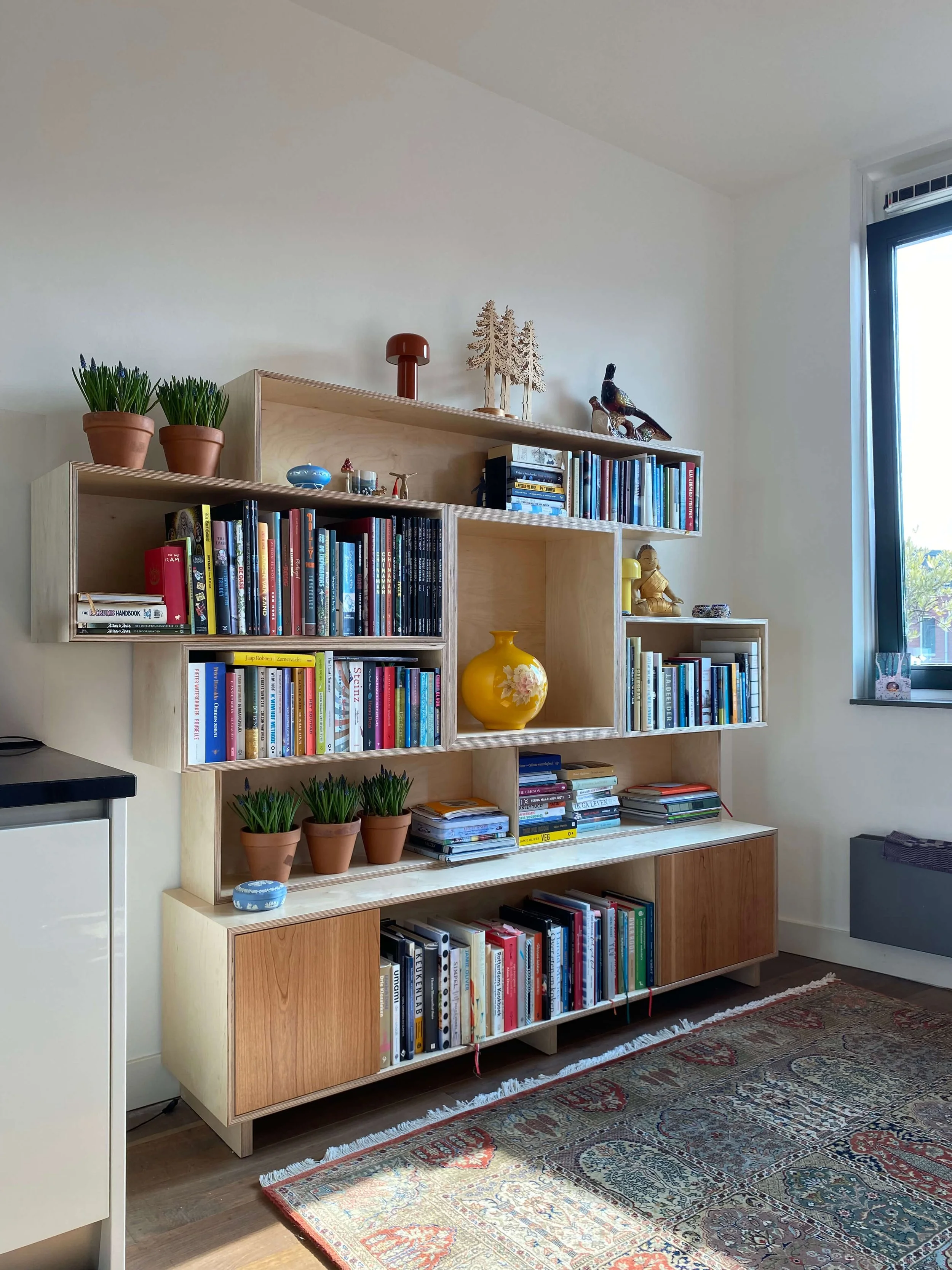 A wooden bookshelf filled with books and decorative items, with potted plants on top and a large yellow vase in the center, positioned in a living room with a window and a patterned rug.