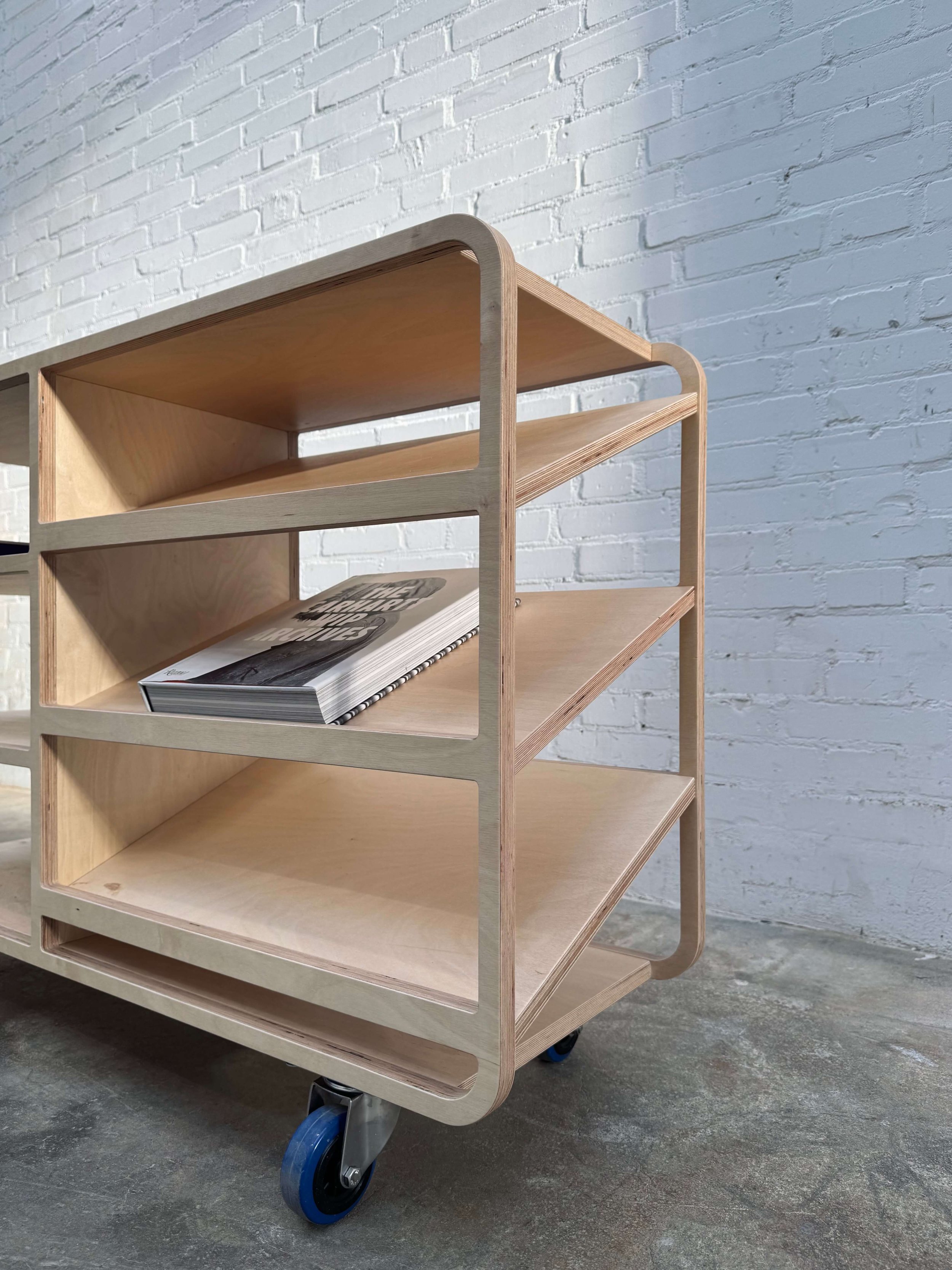 A light wooden rolling cart with three shelves and a curved frame, placed on a concrete floor against a white brick wall. One magazine is on the middle shelf.