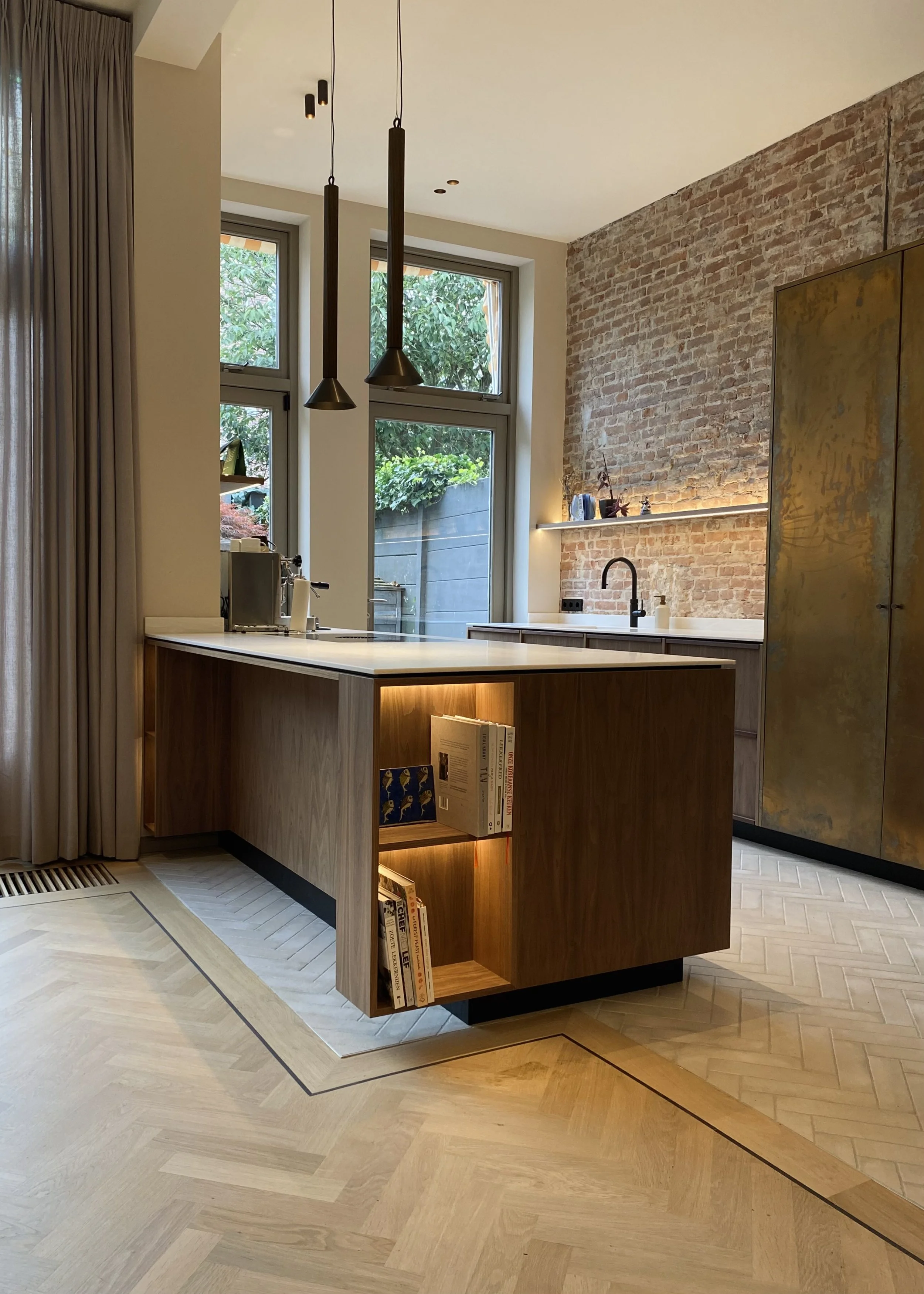 Modern kitchen with a brick accent wall, wooden cabinetry, a kitchen island with built-in bookshelves, pendant lighting, and large windows letting in natural light.