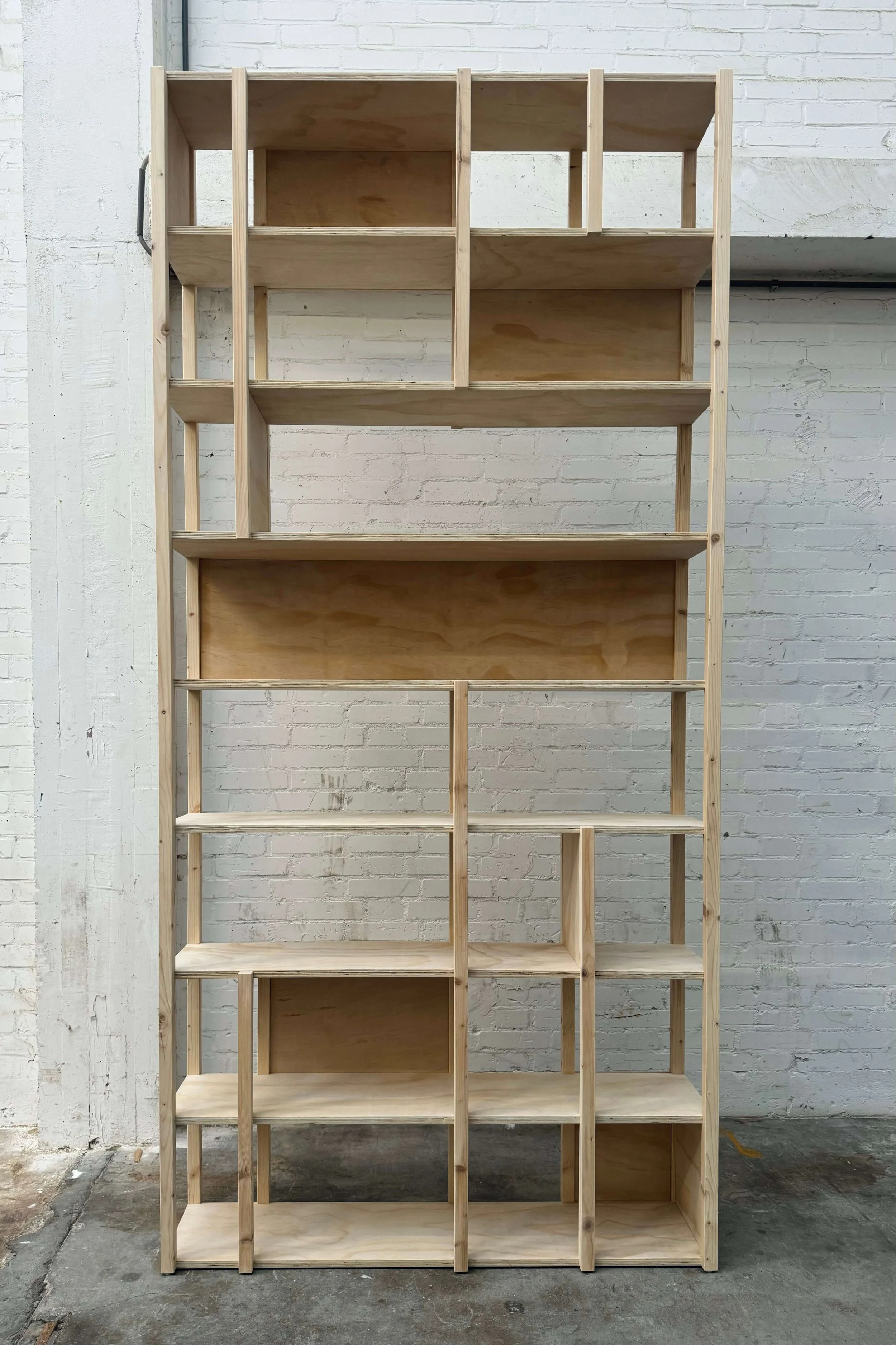 Unfinished wooden shelving unit against a white brick wall in a workshop.