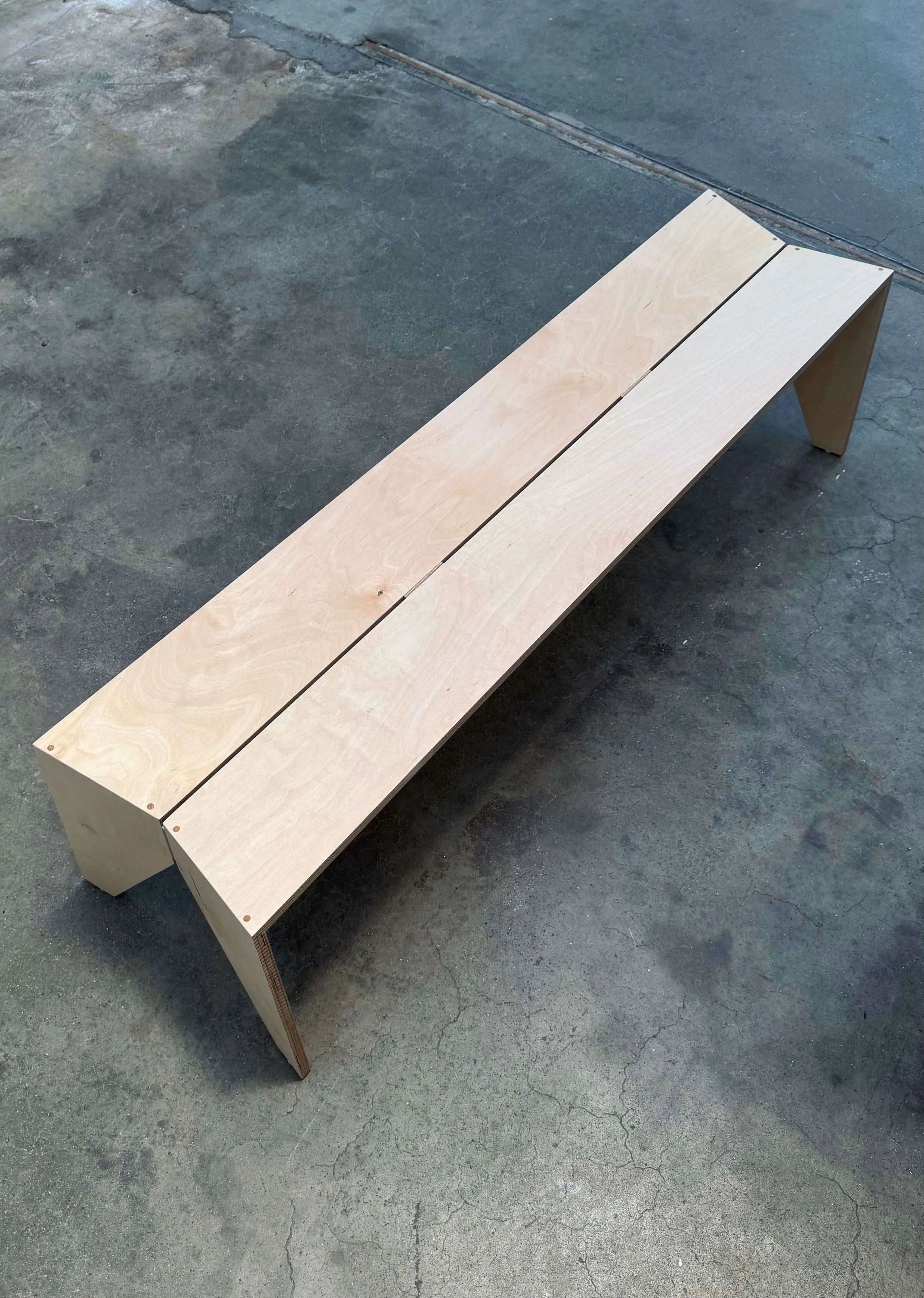Unfinished wooden bench with two planks forming the seat and two angled legs, placed on a concrete floor.