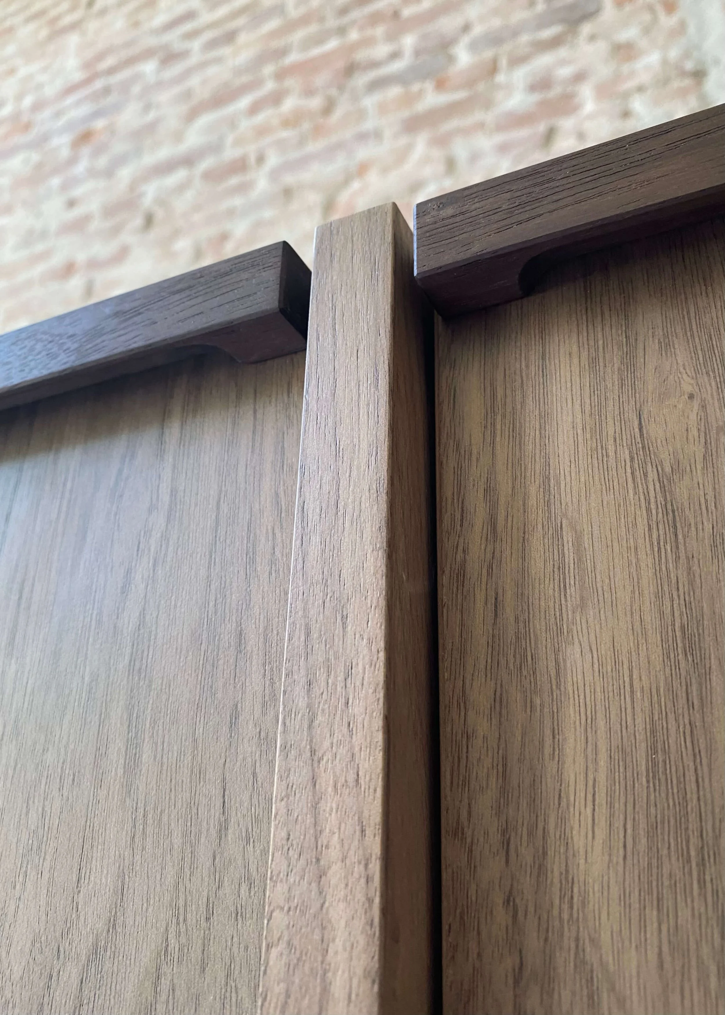 Close-up photo of a wooden cabinet with a brick wall in the background, showing the top corner where two cabinet doors meet.