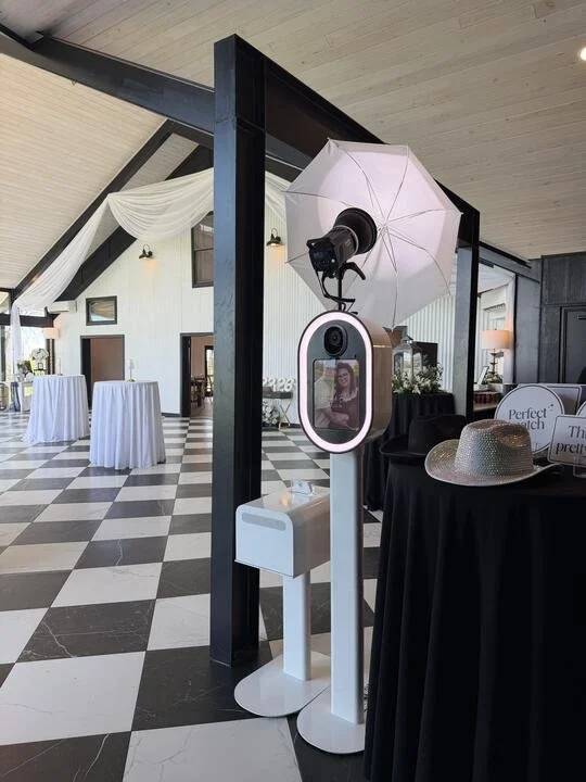 Photo of a photo booth setup in Fort Worth with a DSLR camera, umbrella for lighting, and a mirror, inside a decorated event space with black and white checkered flooring and white draped fabric and props on a table.