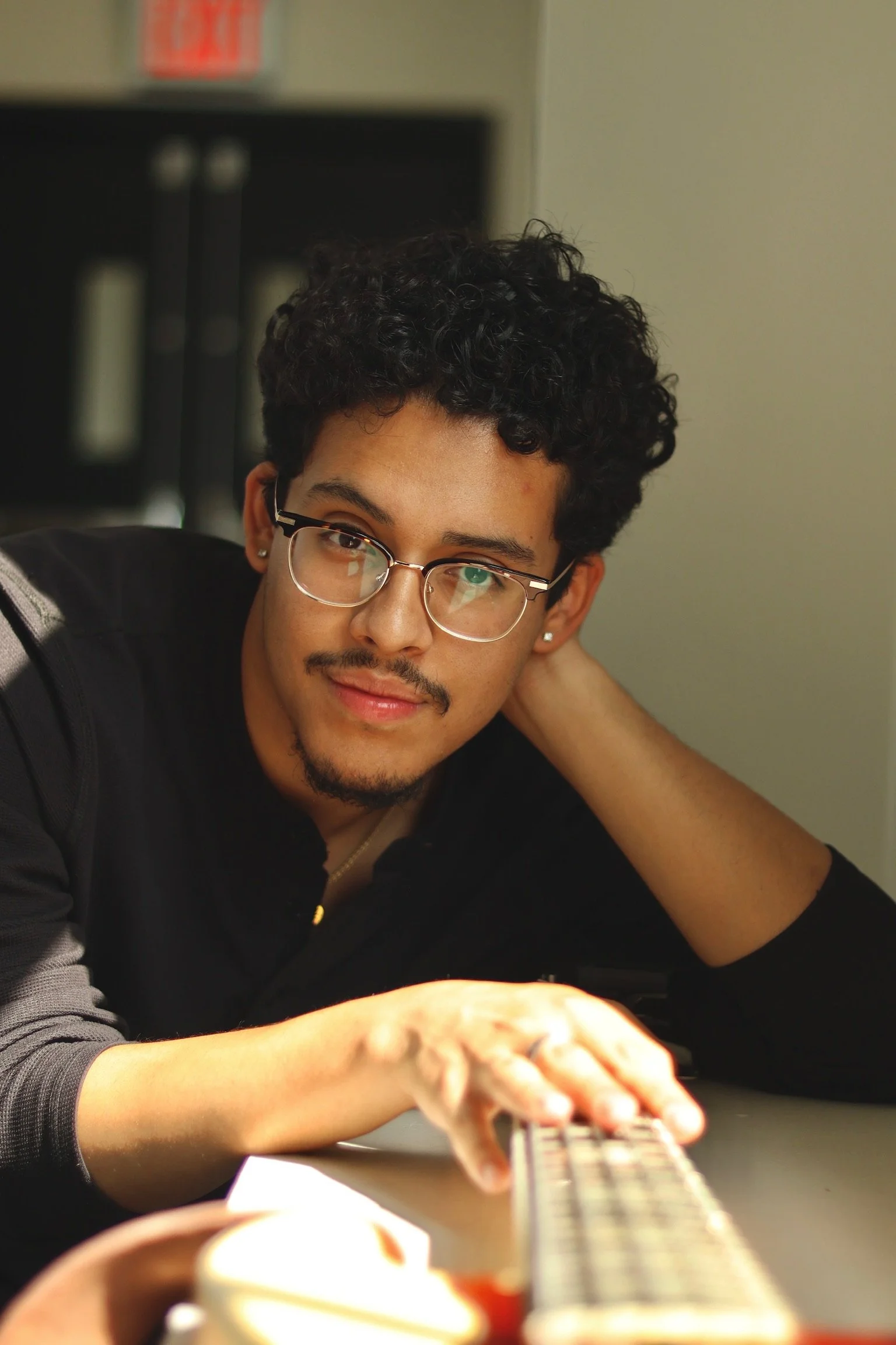 A young man with curly hair, glasses, and earrings, resting his head on his left hand while reaching out with his right hand to play a bass guitar on a desk, with black doors and an exit sign in the background.
