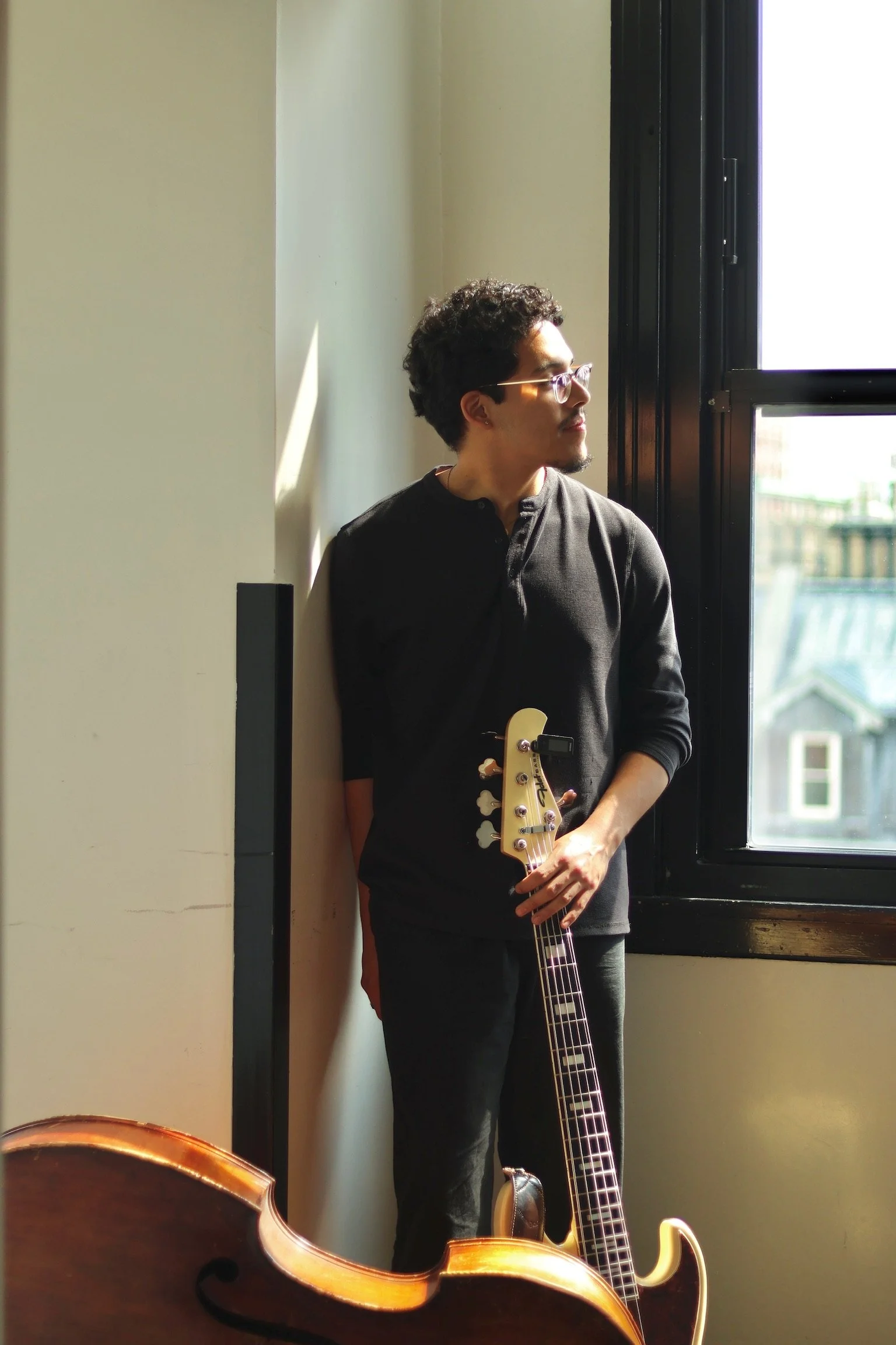 A man standing by a window holding an electric guitar in a room with natural light.