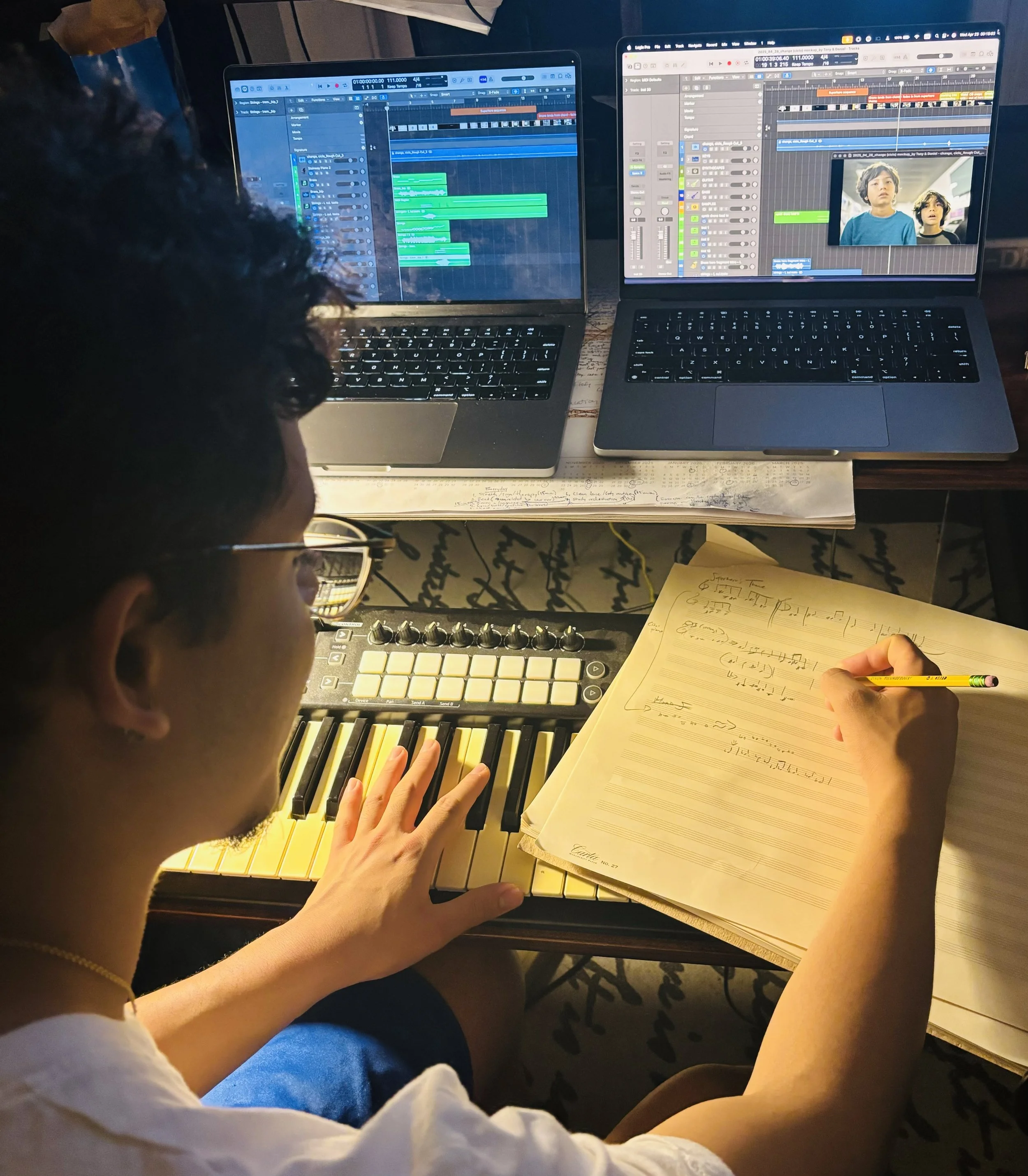 A person working on music production with a keyboard, an MPC pad, two laptops with digital audio workstation software, and handwritten notes.
