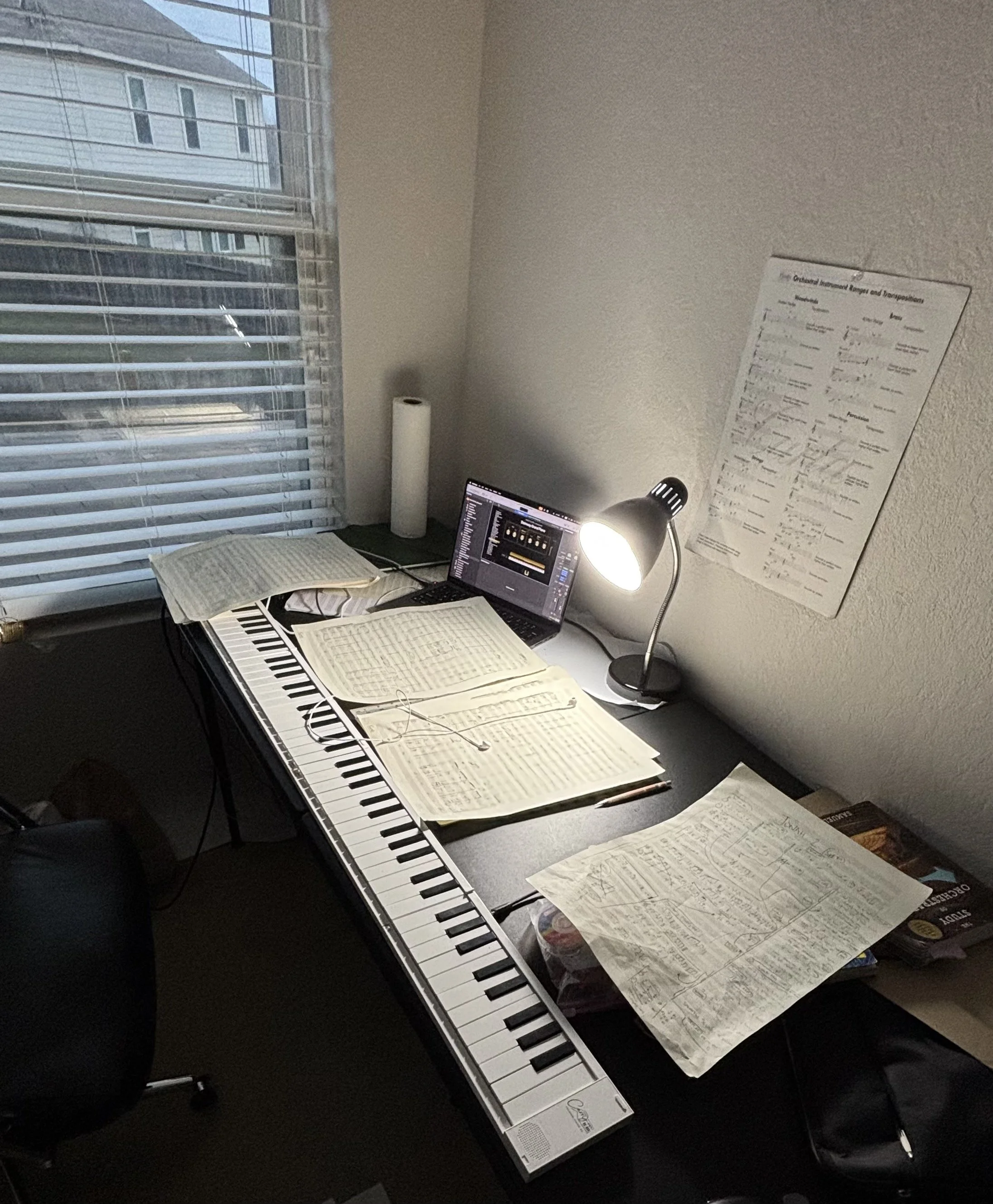 A cluttered desk with a music keyboard, open sheet music, a laptop, a desk lamp shining, a paper towel roll, and a poster on the wall, near a window with blinds.