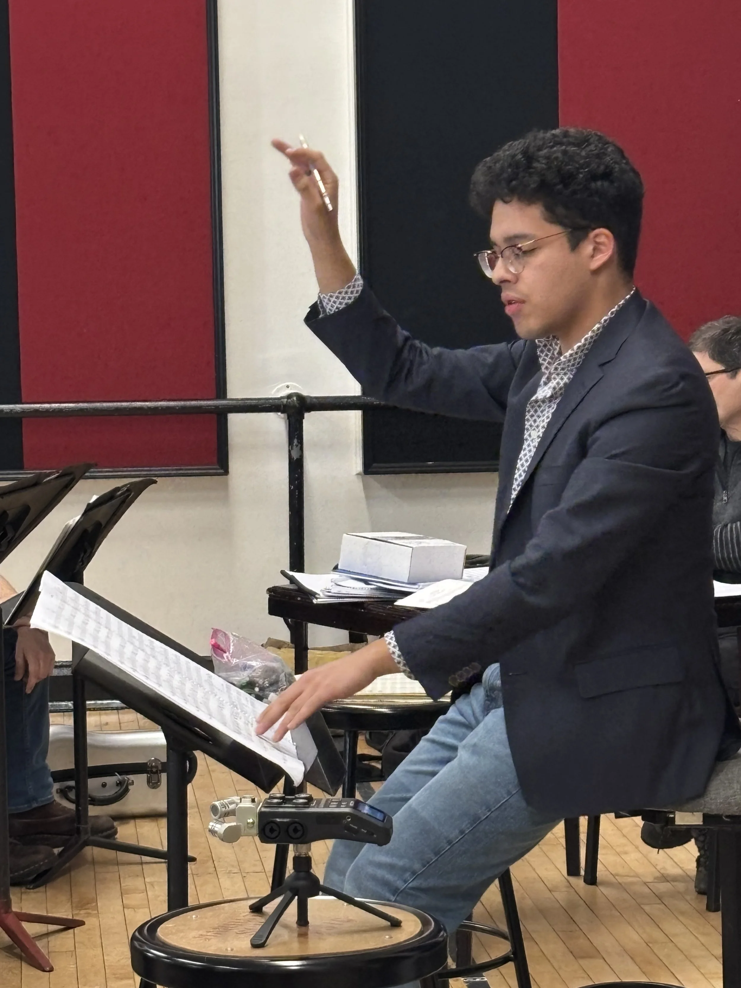 Young man conducting an orchestra, wearing glasses, a dark blazer, and jeans, seated with a music stand, sheet music, and a small percussion instrument, in a rehearsal room with wood floors and red and black wall panels.